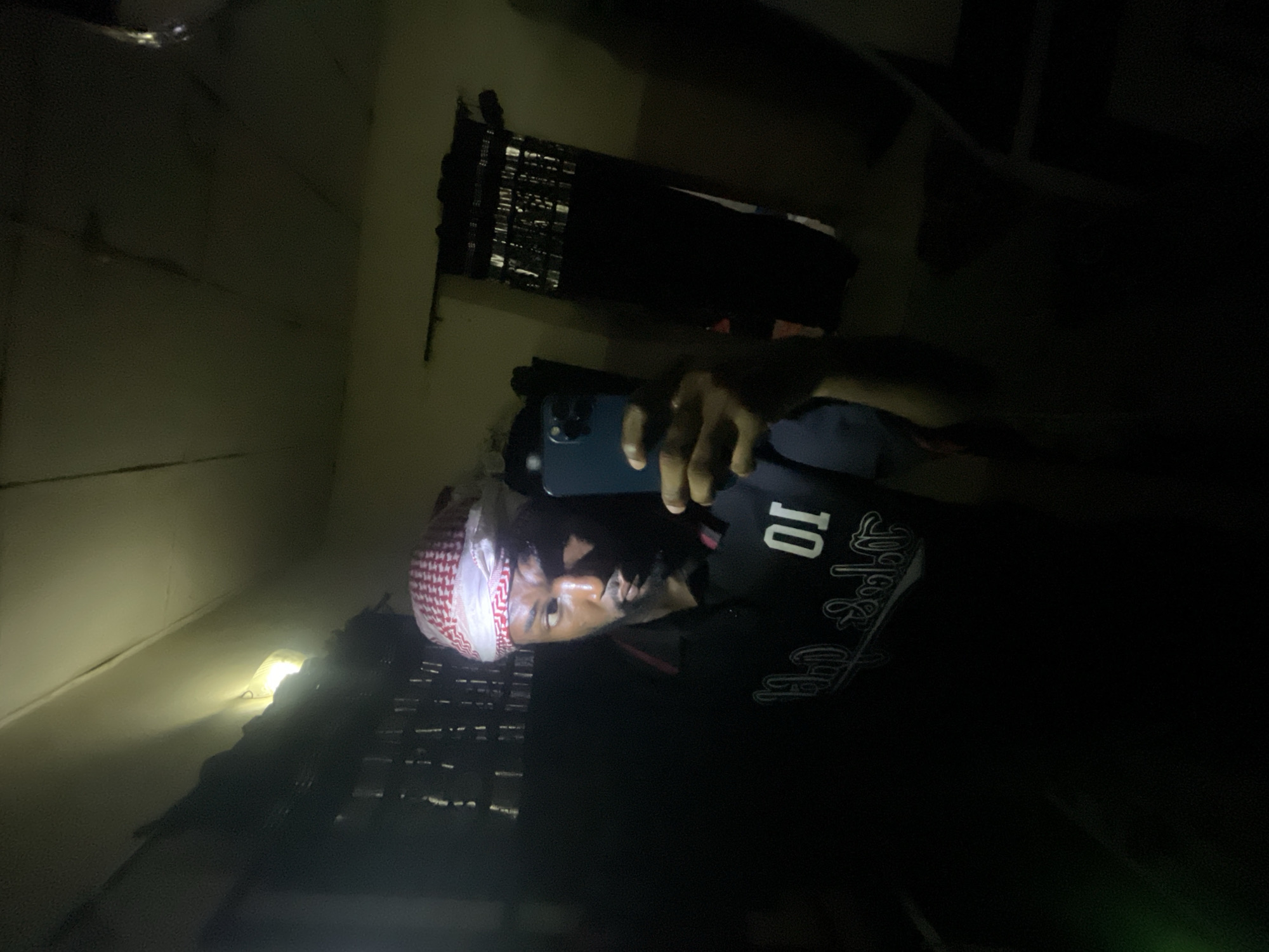 A person wearing a red and white checkered head covering and a black t-shirt is taking a photo with a blue smartphone. The location appears to be indoors, possibly a residence, judging by the wall and the shelf-like structure behind them. The environment is dimly lit. It is not possible to confirm if the location is Lokoja, Nigeria.