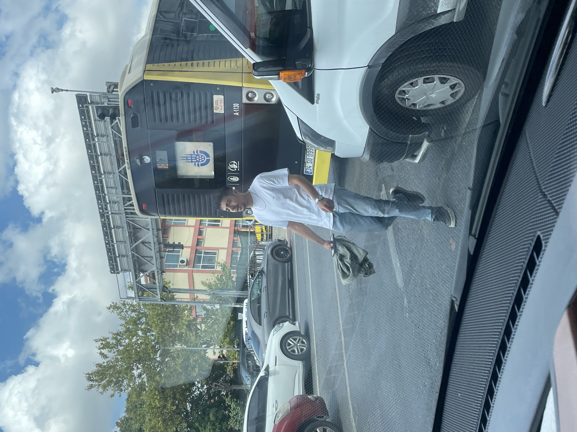 A young man wearing a white t-shirt and blue jeans walks across a city street in Istanbul, Turkey, holding a green cloth in his right hand. He is in motion, looking towards his right. He wears a watch on his left wrist and dark sneakers. Behind him, a yellow and black bus with the Istanbul Metropolitan Municipality logo is stopped at what appears to be an intersection. The bus has license plate "A-1130" and "34 SU 0998". A white van is visible on the right side of the frame, partially obscuring the bus. Several other cars are stopped at the intersection. The sky is blue with scattered clouds. The scene suggests traffic congestion on a sunny day in Istanbul.