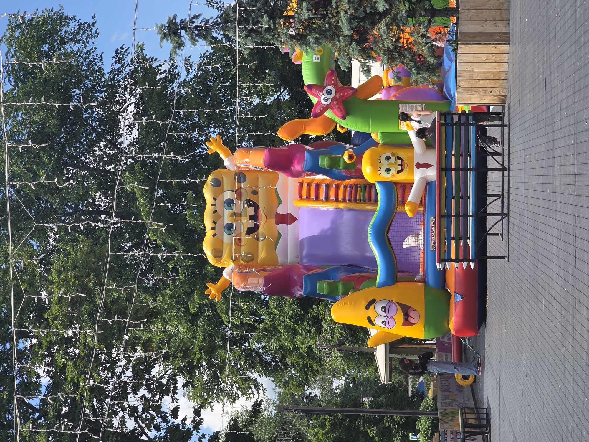An outdoor, daytime scene features a vibrant, inflatable Spongebob-themed playground as the central subject. The inflatable structure is a multi-colored castle with slides and various character figures from the cartoon, including Spongebob Squarepants and Patrick Star. A string of white lights is draped across the top of the frame, above the inflatable. In the foreground, a wooden deck extends into the scene, with a colorful, slatted bench in the mid-ground. Two individuals, a woman and a child, are seated on the bench. To the left of the bench, a young woman in ripped jeans and a dark top is standing and looking at her phone. Behind the inflatable structure, lush green trees and a blue sky are visible, suggesting a park or recreational area. To the right of the inflatable, a pine tree stands tall. The overall atmosphere is cheerful and playful, indicative of a child-friendly entertainment venue.