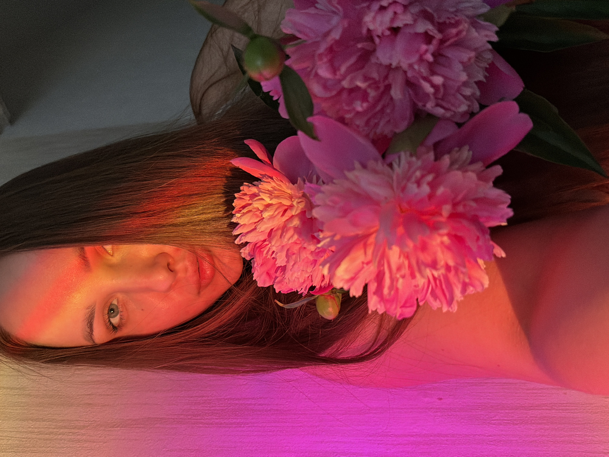 A portrait of a young woman is captured from the chest up, with her face partially illuminated by a pink and orange light, casting a colorful glow across her skin and long, dark hair. She is holding a bouquet of large, pink peonies in front of her. Her left eye is visible, and her lips are slightly parted. The background consists of a textured wall on the left, illuminated by pink light, and a darker, out-of-focus area on the right. The overall impression is one of soft lighting and a delicate, floral subject. There are no discernible elements that place this image specifically in Chaikovsky, Russia.