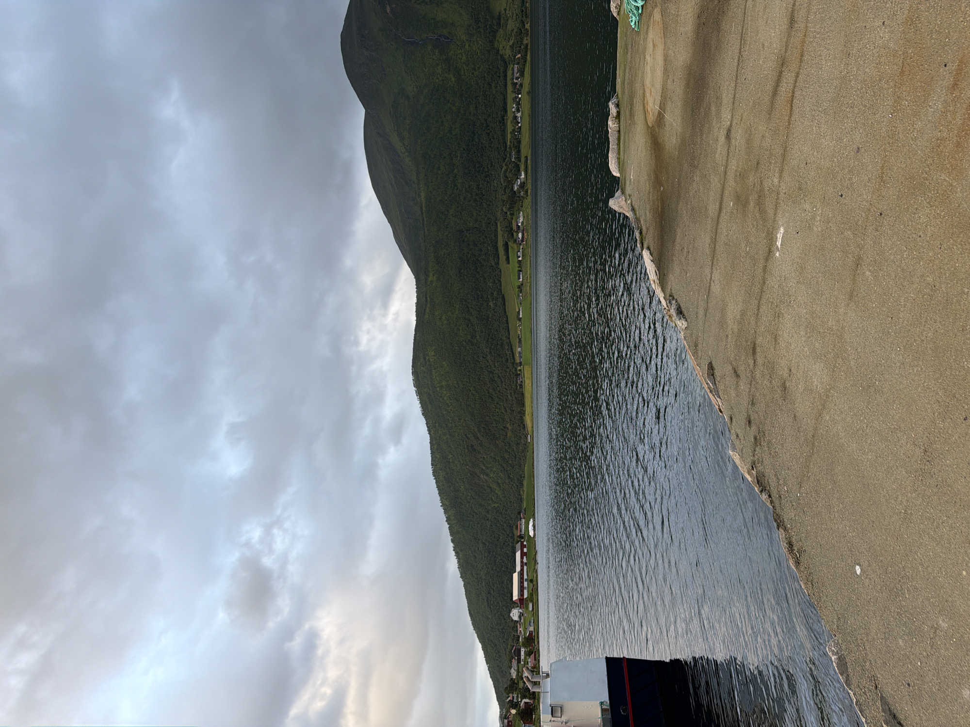 The image depicts a serene view of a body of water, likely a fjord, with a lush, green mountain range in the background. A small village is nestled at the foot of the mountain, with buildings featuring red roofs, suggesting a Nordic aesthetic. The sky is overcast with dramatic clouds, hinting at cooler weather. In the foreground, a concrete dock or pier extends into the water, with a portion of a boat visible on the left side. The water is calm, with gentle ripples reflecting the muted sky. The overall impression is one of natural beauty and tranquility, characteristic of a coastal Norwegian landscape. The location is the city of Harstad, Norway.