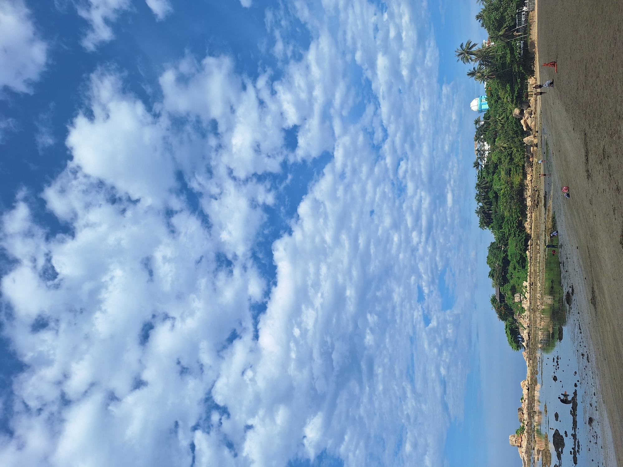 The image captures a serene beach scene in Nha Trang, Vietnam. The sky is a vibrant blue, dotted with scattered white clouds, suggesting fair weather. The composition is dominated by the expansive sky on the left and the sandy beach and coastline on the right.
Along the shoreline, lush green trees and palm trees create a tropical backdrop, interspersed with large rocks. A distinctive white and blue dome-shaped structure is visible amidst the greenery, hinting at a resort or a local landmark. The beach itself is a mix of sand and shallow water, reflecting the sky and surrounding vegetation. Scattered along the beach and in the water are a few individuals, some walking or standing, and one person appears to be sitting near the water's edge. The scene evokes a sense of tranquility and leisure, likely during the daytime. The lighting suggests it could be morning or late afternoon, with a soft, diffused light. The overall impression is one of a beautiful and peaceful coastal environment.