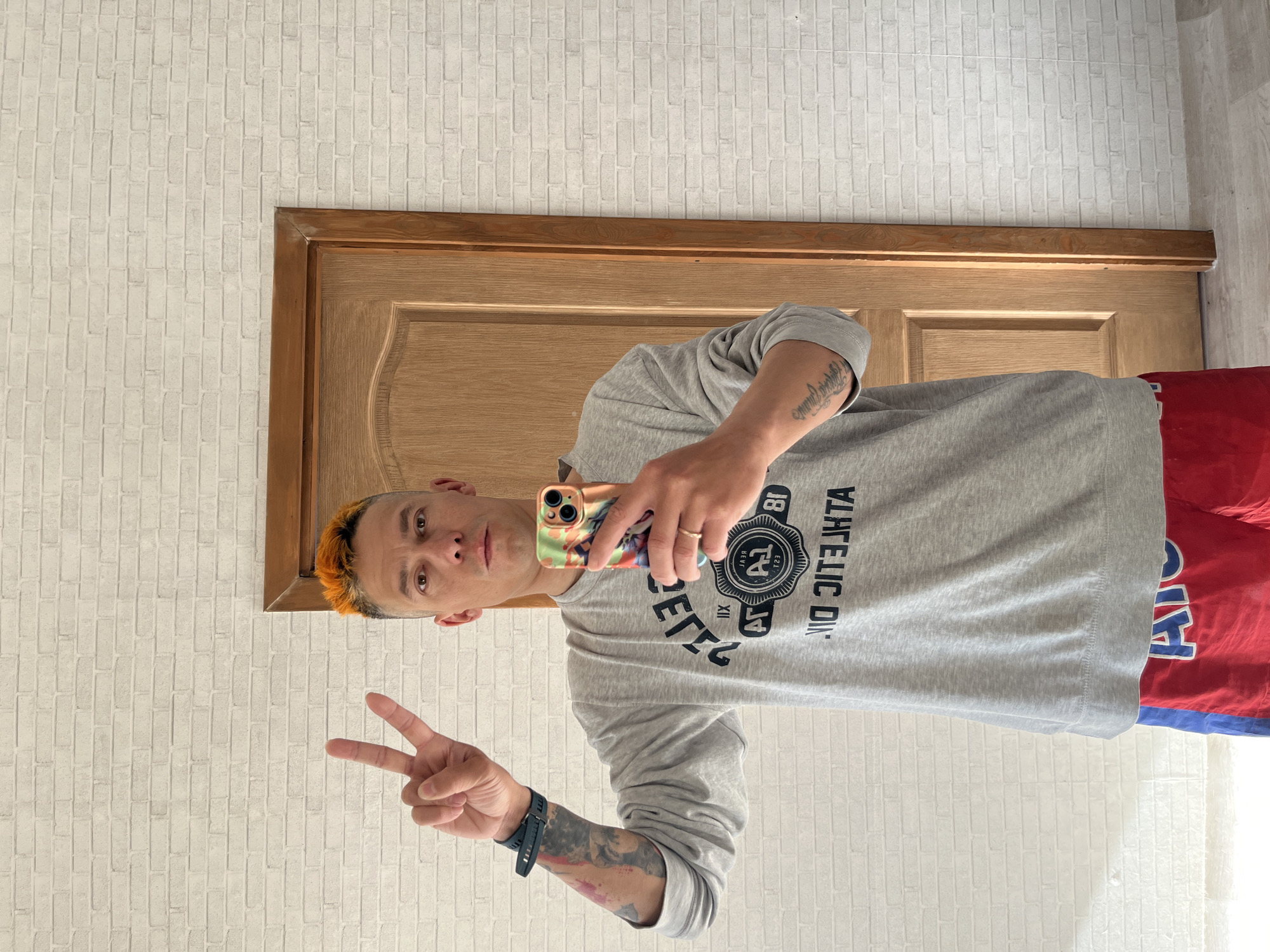 A man with orange hair is taking a selfie with a phone in front of a closed wooden door. He is wearing a gray long-sleeved shirt with a black design, a wristwatch, and red shorts. He is making a peace sign with his left hand. The wall behind him has a white brick pattern. The location is implied by the request to be in Chaikovsky, Russia.