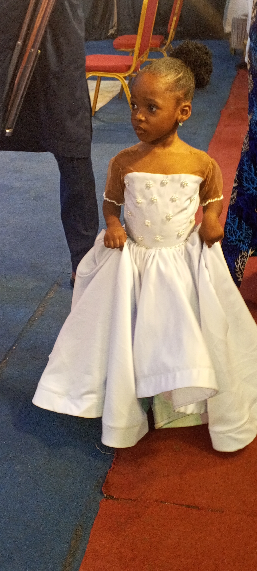 A young girl, appearing to be a flower girl, stands in a white dress with floral embellishments on the bodice. The dress has a gathered skirt that reaches the floor. The girl has dark hair styled in a bun and wears small earrings. She is looking to her right.
In the background, there are chair backs and legs, and a portion of someone's leg is visible to the left. A blue and red patterned carpet is on the floor. The picture was taken at Sabon Tasha, Nigeria.