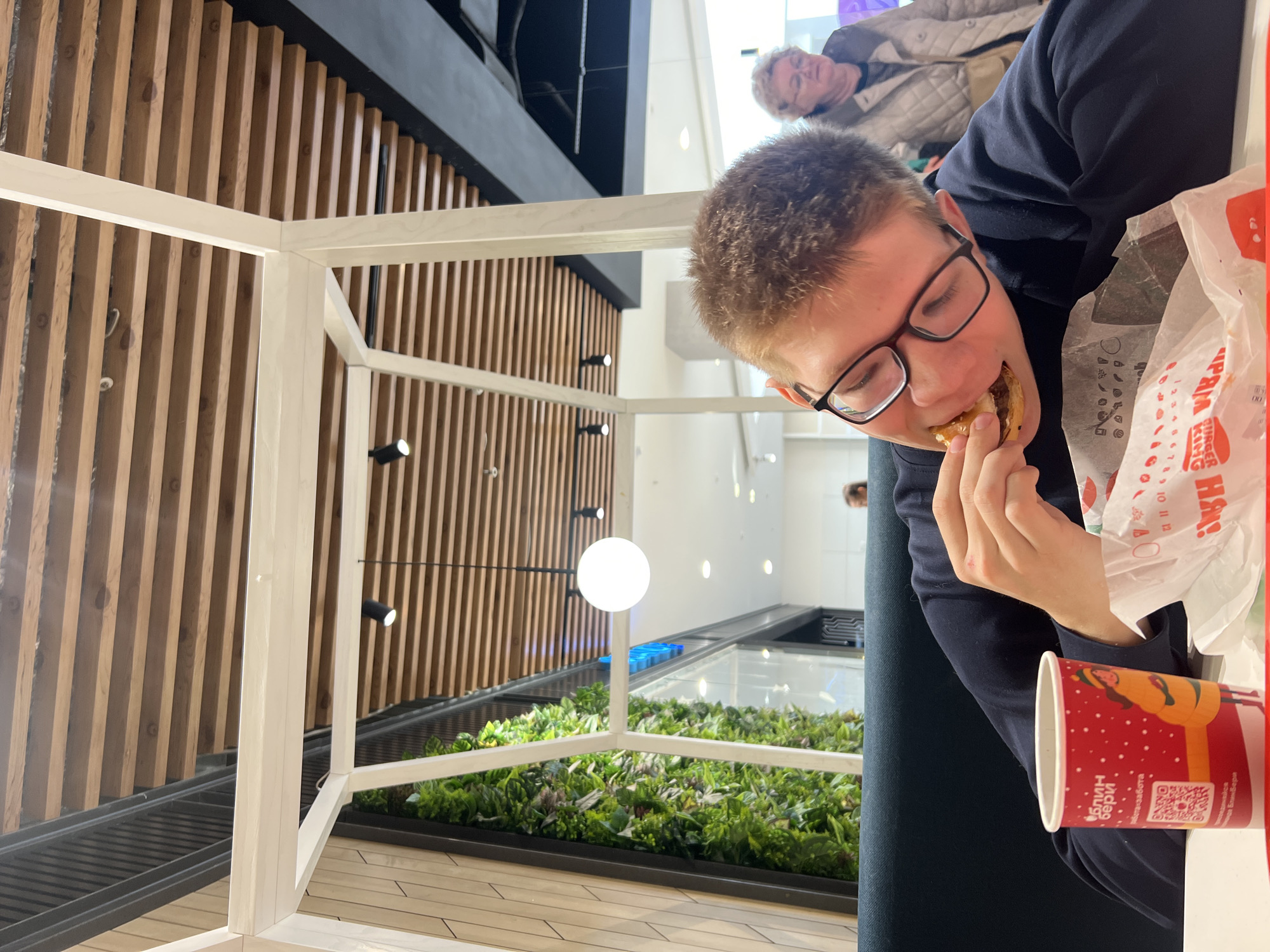 The scene is set within the food court of TRC Talisman shopping mall, located at Kholmogorova Street, 11, Izhevsk, Russia.
A young male, appearing to be in his late teens or early twenties, is seated at a white table in the foreground. He has short, light brown hair and wears black-rimmed glasses and a dark blue long-sleeved shirt. He is actively consuming a burger, holding it with his right hand and taking a bite. On the table in front of him, a crumpled white paper wrapper, bearing the "BURGER KING" logo in Cyrillic, holds the remainder of the food item. To his left, a red paper cup with a festive illustration, a QR code, and Cyrillic text indicating "БлинБерри" (BlinBerry) is also on the table.
In the blurred background, to the right of the main subject, an older woman with short, light-colored, curly hair and glasses is visible, wearing a light-colored quilted jacket as she appears to walk past.
The environment features a modern interior design. The ceiling is characterized by parallel wooden slats, interspersed with black track lighting fixtures and a prominent spherical white pendant light. A decorative white, open-frame structure defines the immediate seating area. To the left, a large vertical panel is covered with artificial green foliage, creating a "vertical garden" effect. The seating appears to be upholstered booths in a dark green or teal color. The background shows hints of other mall sections, including glass partitions and distant white walls.