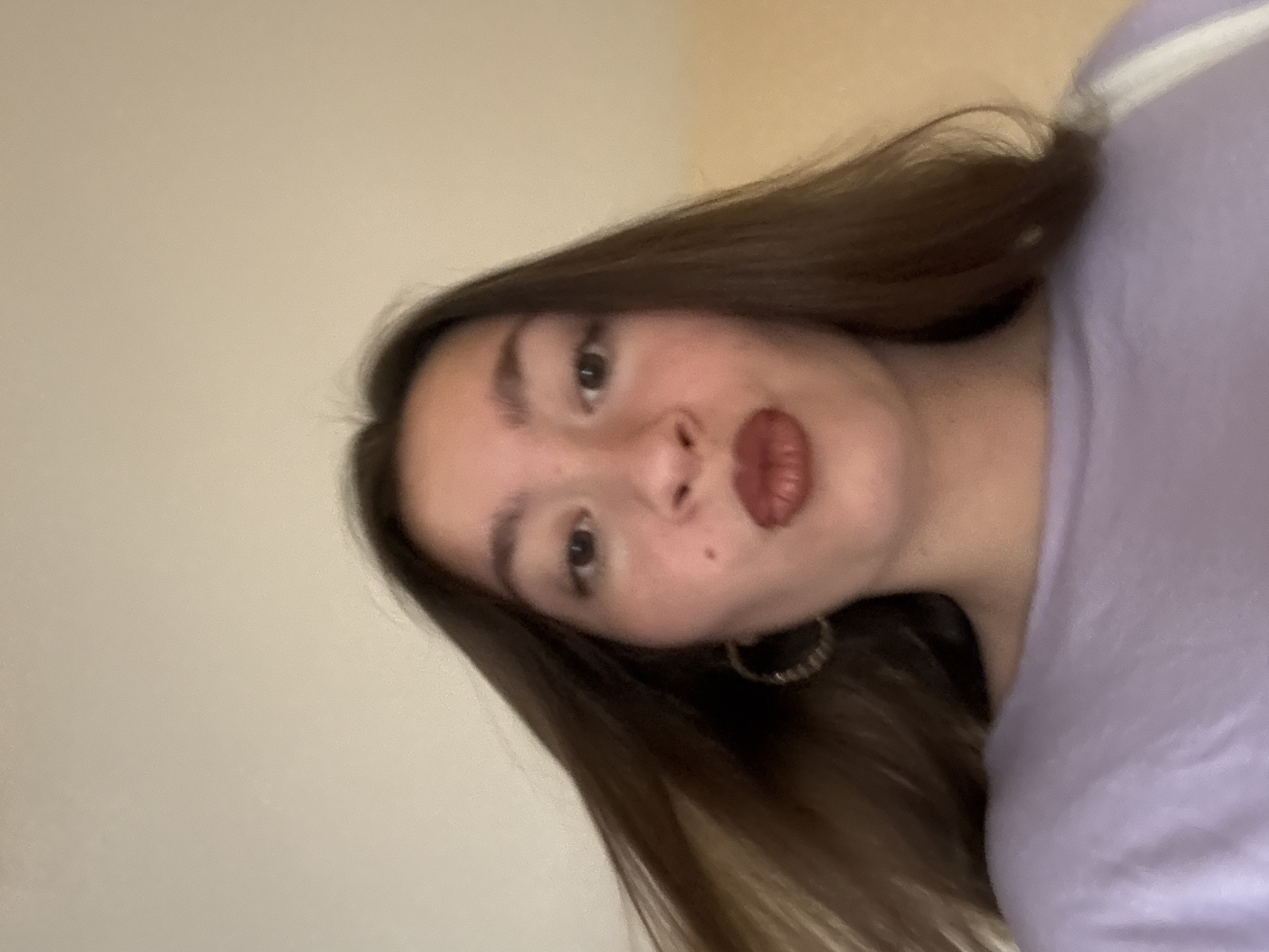 An image shows a close-up, low-angle selfie of a young woman with brown hair, wearing a light purple top and hoop earrings. She is pouting her lips, which are adorned with dark lipstick. Her hair is partially obscuring her face, with strands blowing to the left. The background is a plain, light-colored wall, suggesting an indoor setting. No specific location in Sofia, Bulgaria can be identified from the visual information present.
