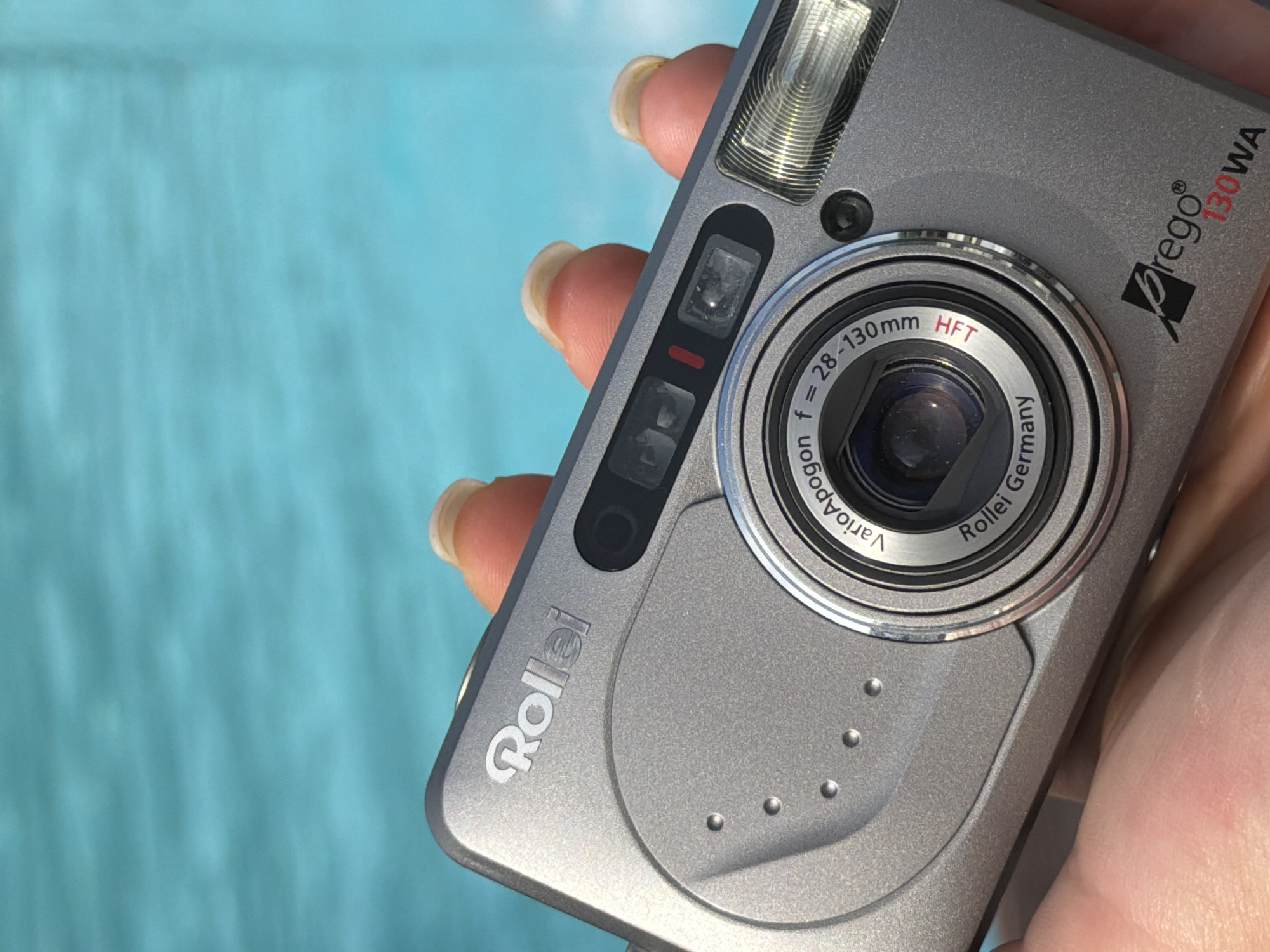 A close-up, high-angle shot captures a person's hand holding a silver Rollei Prego 130WA compact film camera against a blurred light blue background, suggestive of water, likely a swimming pool or the Black Sea near Varna, Bulgaria. The camera is held diagonally, showcasing its front, top, and part of its left side.
The camera's metallic grey body features a pop-up flash unit at the top, and below it, an optical viewfinder window and a red light. The prominent lens barrel dominates the center, with visible text "VarioApogon f = 28-130mm HFT Rollei Germany" encircling the glass. The "HFT" text is in red. To the left of the lens, the "Rollei" brand logo is embossed, and to the right, the model name "Prego® 130WA" is printed, with "130WA" in red lettering. The camera's surface has a slightly textured finish.
The hand, with light skin and neatly manicured fingernails, wraps around the camera's side and bottom. The background's soft blue hue and subtle ripples create a serene and summery setting, indicating bright daylight conditions. The image focuses sharply on the camera and hand, with the background softly diffused.