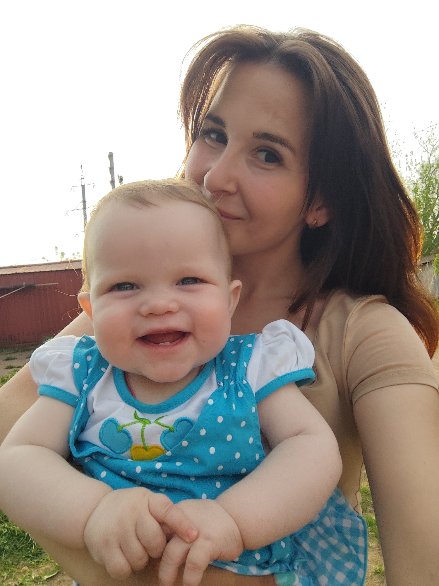 A woman with brown hair and a light complexion is holding a baby. The baby is wearing a blue and white polka-dot dress with a cherry design on the front. The woman is wearing a tan t-shirt. The background appears to be a grassy area with a red building and power lines visible. The image was captured outdoors and includes a hint of Фоки, Russia.
