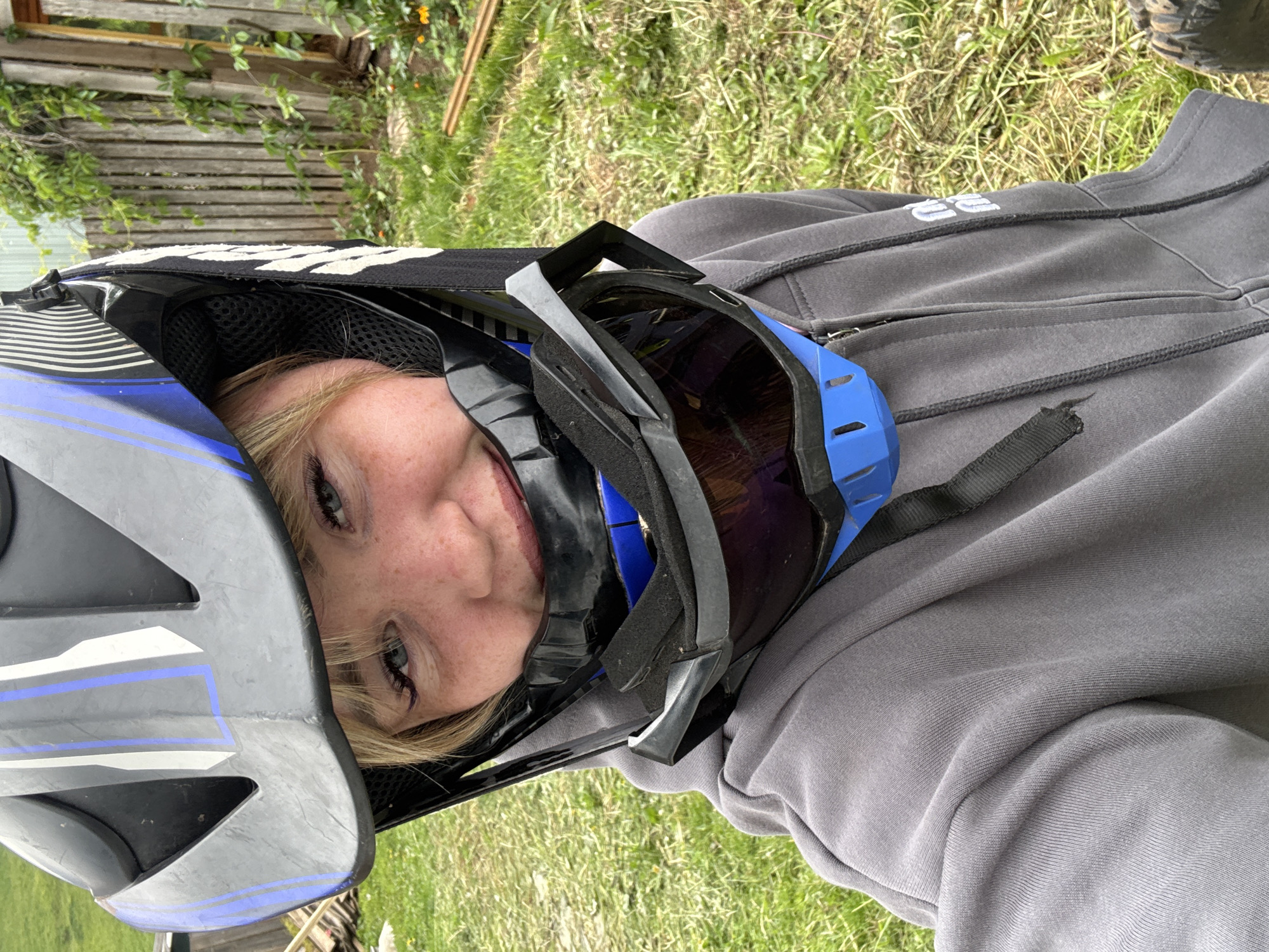 An outdoor, eye-level photograph taken in a selfie style captures a young individual in Ляльшур, Russia. The subject, appearing to be female with blonde hair and visible freckles on the cheeks, smiles subtly while looking directly at the camera. They are wearing a dark grey full-face helmet adorned with blue and white vertical stripe accents. A pair of black-framed goggles with dark, reflective lenses is positioned around the neck, beneath the helmet's chin guard. The subject is dressed in a dark grey full-zip hoodie with ribbed cuffs, displaying a white "NO ME" logo on the right sleeve.
The background depicts a grassy outdoor area. To the right of the subject, a weathered vertical plank wooden fence is visible, partially covered by green climbing foliage and small orange flowers at its base. Behind the fence, a light-colored building structure is present. On the ground to the right, several pieces of lumber or wood debris are scattered amongst the grass. The lighting suggests natural daylight.