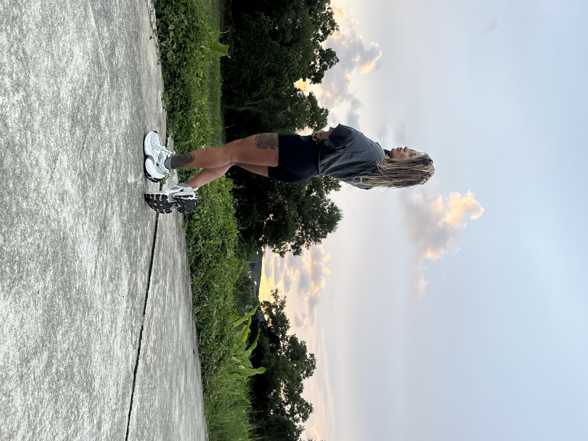 This media file shows a young woman standing outdoors on a concrete path. She is wearing athletic shorts and a grey t-shirt. Her hair is long and blonde. She has tattoos on her legs. She is wearing white and black athletic shoes. The woman is looking upwards and to the right. Behind her is a line of trees and lush green vegetation. The sky is a pale blue with some soft, fluffy clouds, and the light suggests it is either sunrise or sunset, casting a warm glow on the clouds. The scene appears to be set in a rural or suburban area with a natural landscape. The overall mood is calm and serene. No text is visible in the image. The location context provided is Thep Krasatti, Thailand.