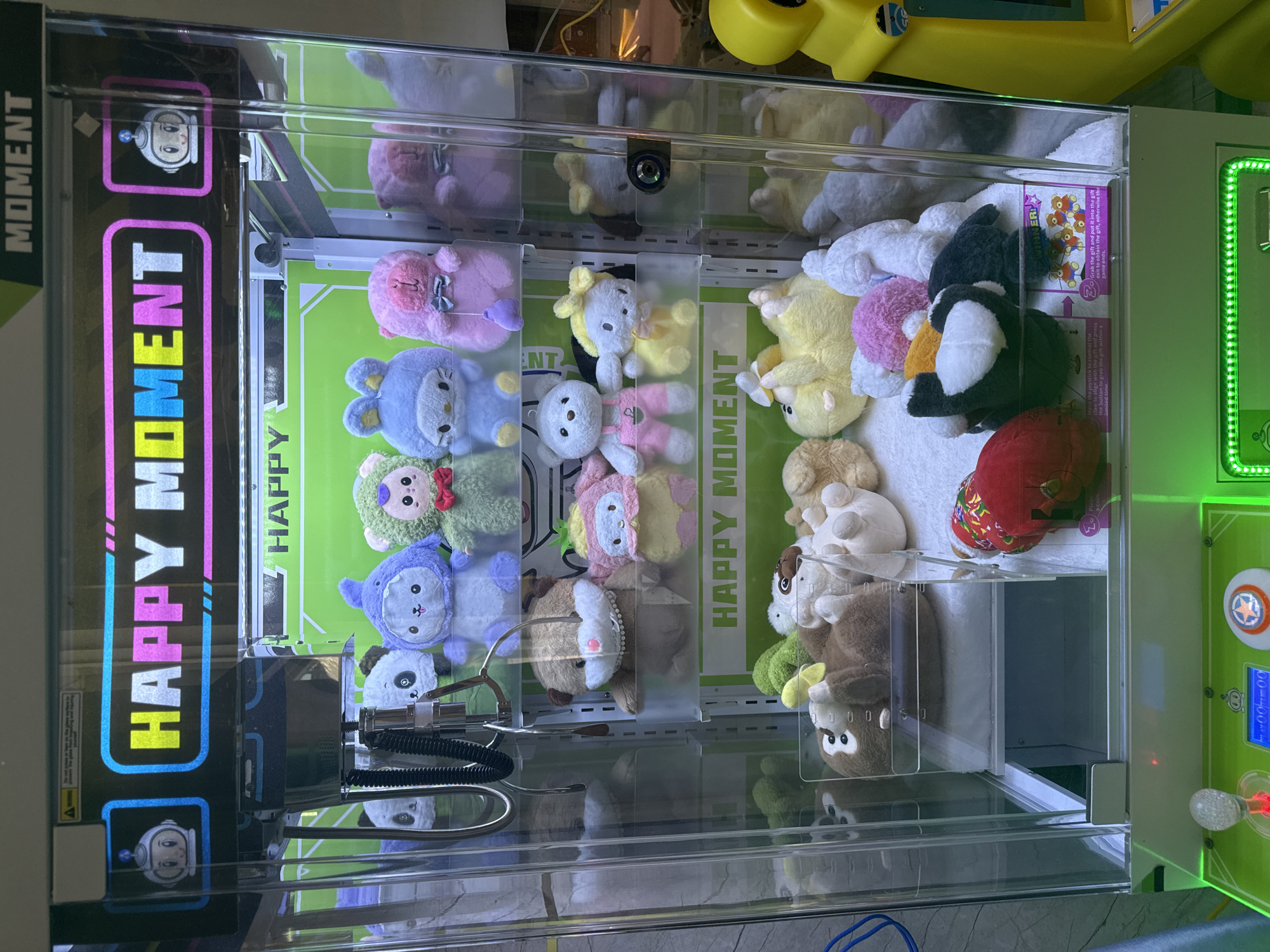 This is a medium shot, eye-level, full-frame photograph of a claw machine filled with plush toys, located in a commercial establishment in Bac Nha Trang Ward, Vietnam. The machine is predominantly clear plastic with a green and white interior. The top of the machine features a bright LED-lit sign with "HAPPY MOMENT" emblazoned across it, flanked by blue and white robot icons. Inside the machine, various plush toys are arranged on three shelves. The top shelf displays blue, purple, and green animal-shaped plush toys, some with accessories like bows. The middle shelf holds brown, pink, and white plush toys, including teddy bears and a rabbit character. The bottom shelf contains more plush toys, including a brown monkey, a yellow chick, and other assorted characters. The prizes are displayed on a white, carpeted surface within the machine. The bottom of the machine has a control panel with a joystick, a digital display, and illuminated green LED lights. The overall scene is brightly lit, and the reflections on the plastic suggest an indoor environment with artificial lighting. There are no people visible in the image.