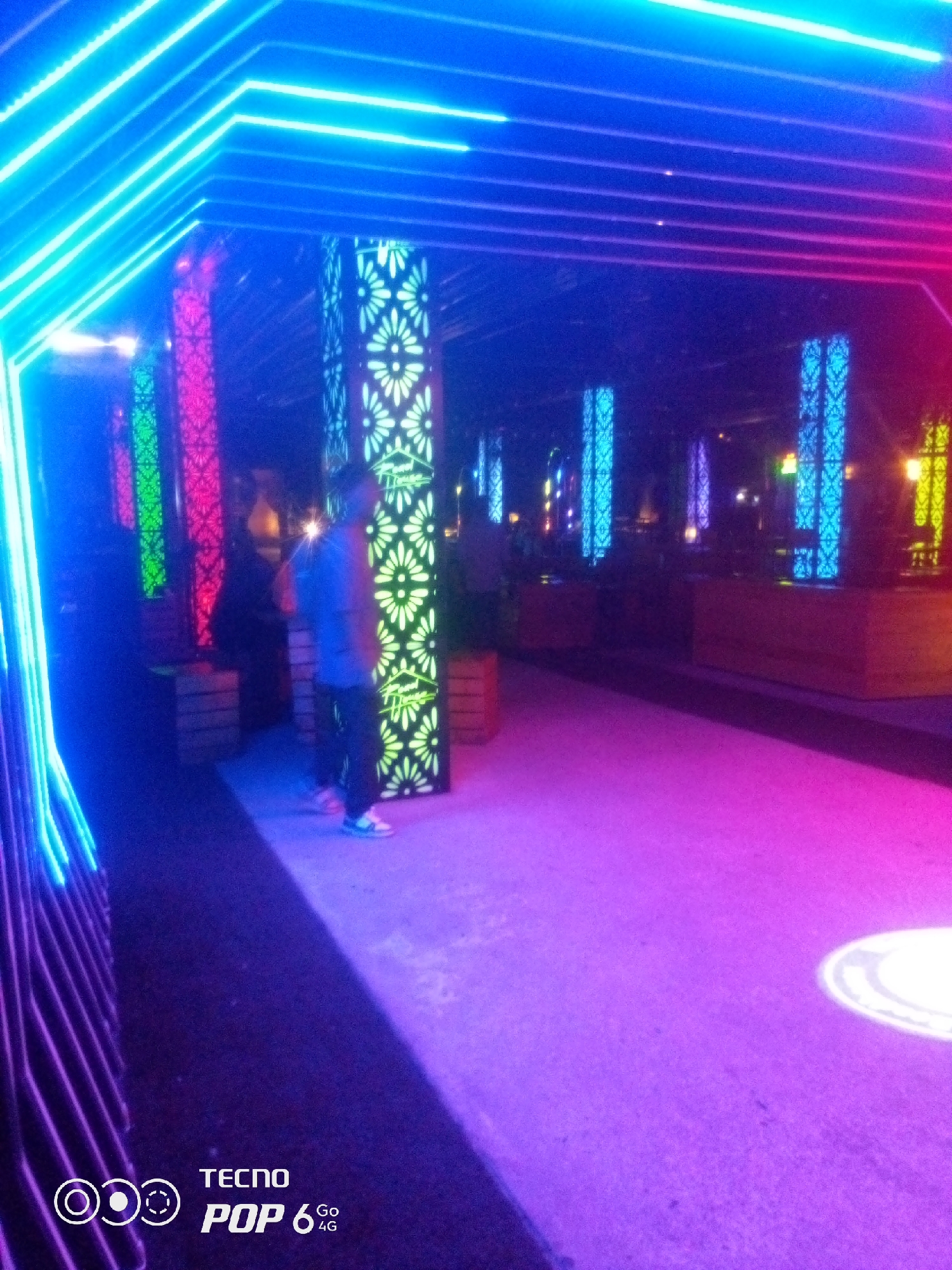 The image shows an outdoor area decorated with vibrant neon lights. The lights are arranged in a geometric pattern, forming a tunnel-like structure with alternating colors of blue, pink, and purple. Several illuminated pillars with intricate floral cutouts are positioned along the sides. In the foreground, a person is standing, facing away from the camera, wearing a blue shirt and dark pants. The ground is covered in a light-colored surface illuminated by the colorful lights, creating a surreal atmosphere. The background is dark, suggesting it is nighttime. The image also features a watermark at the bottom left corner with the text "TECNO POP 6 Go 4G". This scene is located in the city of Howayi, Nigeria.