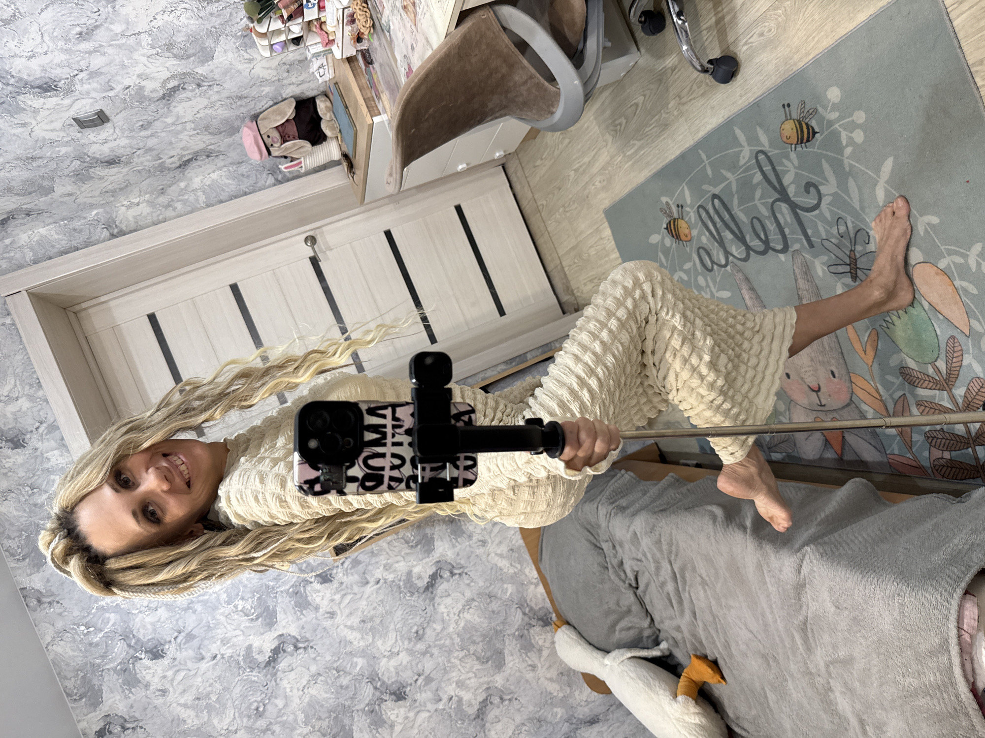 A woman with long blonde hair poses for a selfie using a phone attached to a selfie stick. She is wearing a textured, light-colored outfit. The background includes a light grey and white patterned wall, a light-colored wooden door, and a desk with an office chair. There is also a bed with a grey blanket and a stuffed goose. A rug with a cartoon rabbit and bee design is visible on the floor. The location is Заволжье, Russia.