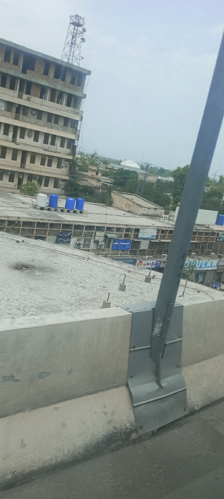 The image shows a view from a road, looking towards a multi-story building under construction, located in the city of Alam Khan Kalay, Pakistan. The building is made of concrete and has a telecommunications tower on its roof. Several blue water tanks are visible on a lower rooftop in front of the building. Signs with Arabic script are attached to the lower building. The foreground is dominated by a concrete barrier and a metal pole. The background shows more buildings, trees, and a domed structure, possibly a mosque. The sky is overcast.