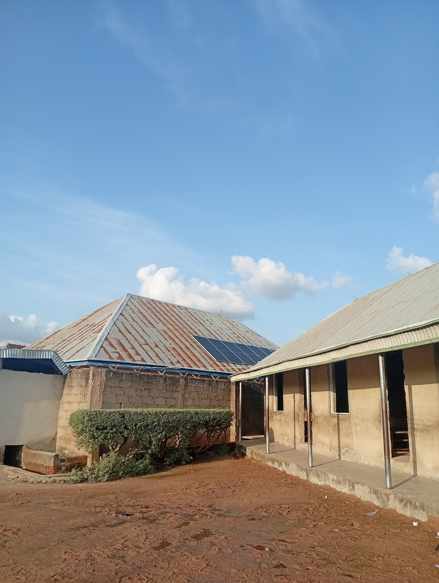 The image depicts an outdoor scene with two buildings in the foreground and a clear blue sky with scattered white clouds overhead, suggesting it is daytime and the weather is fair. The buildings appear to be of simple construction. The building on the left has a gabled roof made of corrugated metal that shows signs of rust and age. Several solar panels are visible on this roof. The walls of this building are made of concrete blocks. In front of this building, a neatly trimmed green hedge runs along the wall.
To the right, another building has a flat or gently sloping roof, also made of corrugated metal. This structure has an open-air walkway or veranda supported by metal posts. The walls of this building are rendered in a light color, and there are several openings that appear to be windows or doorways, some of which lead into dark interiors. The ground in front of the buildings is a reddish-brown dirt, wet in places, possibly from recent rain. Some debris, including a plastic bottle, is visible on the ground.
The scene appears to be in a rural or semi-rural area, possibly a school or community center, given the construction style and the presence of solar panels. There is no visible text in the image. The overall impression is one of functional, perhaps developing, infrastructure under a bright sky.
