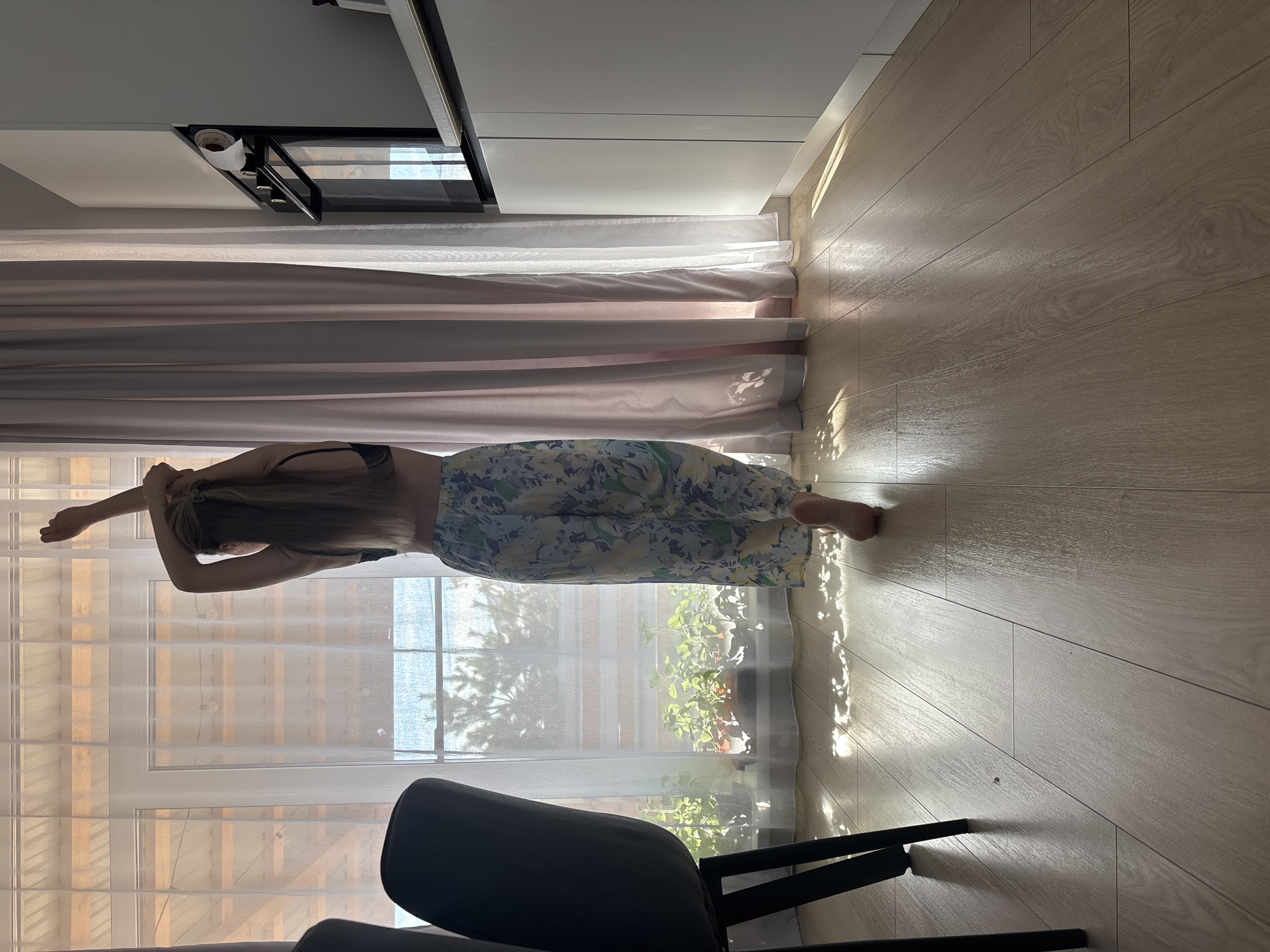 A woman with long blonde hair, wearing a black bra and floral pants, stands in front of a window with sheer curtains. The location is identified as Chaikovsky, Russia. The window looks out onto greenery. Light streams into the room, highlighting wood flooring. A black chair is visible in the foreground, positioned slightly in front of a sofa. A kitchen counter and cabinets are positioned on the left of the image.