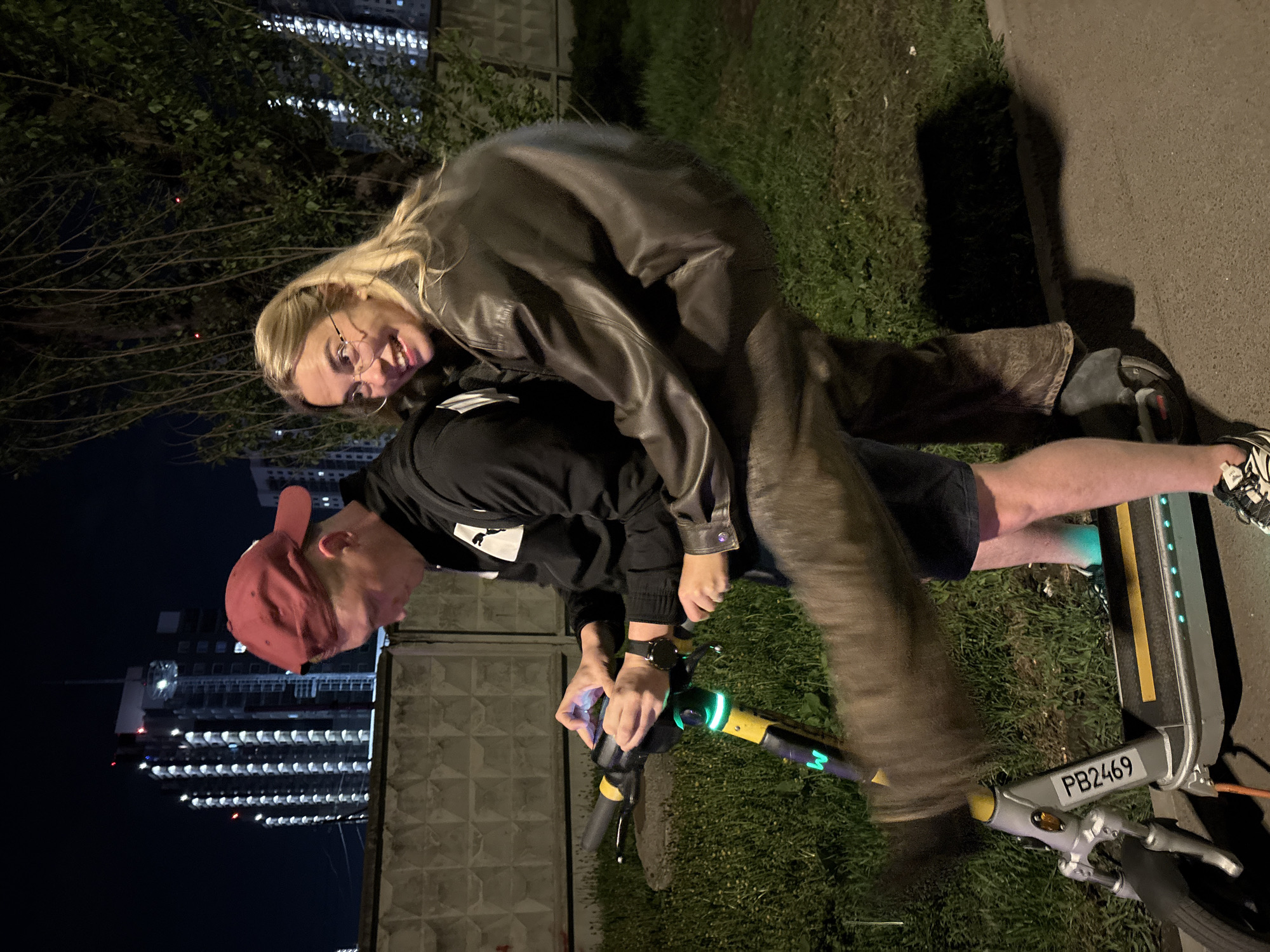 A photo taken at night depicts a man and a woman on an electric scooter with license plate PB2469. The man wears a red baseball cap, black hoodie, and dark shorts. He is operating the scooter while looking down. The woman, positioned behind the man, wears glasses and a leather jacket and appears to be smiling at the camera. The background includes green grass, a concrete fence, and a high-rise building with many illuminated windows at night. This is consistent with the city of Krasnoyarsk, Russia.
