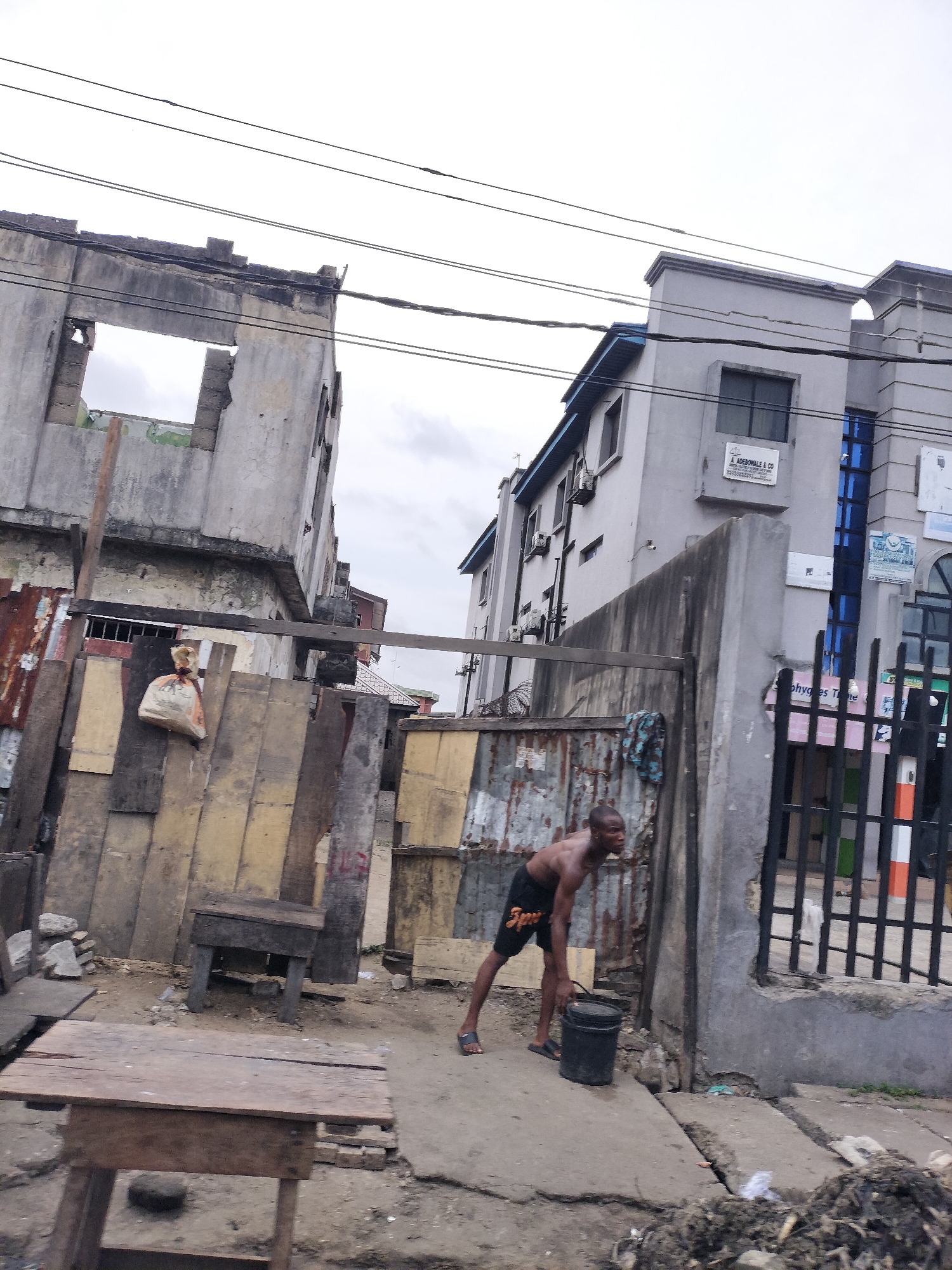 The image depicts a scene in Alaiyabiagba, Nigeria, characterized by contrasting levels of development. A shirtless man in black shorts is carrying a black bucket near a concrete structure, likely a fence or wall, adjacent to a building with commercial signage, a black fence, and a painted orange bollard. Debris is visible on the ground. In the background, there is a dilapidated building lacking a roof and window frames, along with structures constructed of scavenged materials like corrugated metal and wood planks. A cloth sack hangs on one of the wooden structures. Overhead are numerous electrical wires stretching across the frame. The scene suggests a mix of urban decay and established commercial activity.