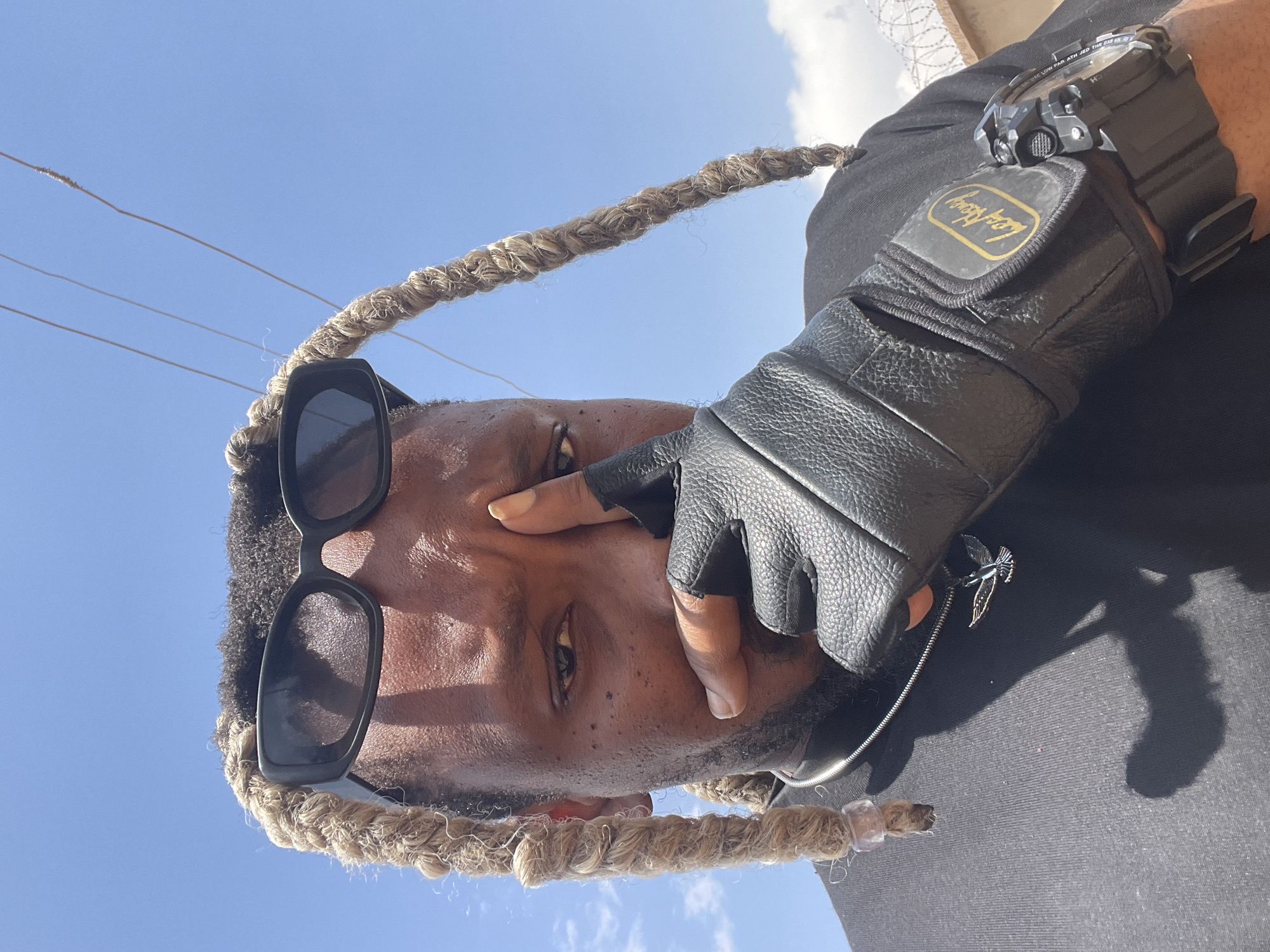 An upward-facing shot shows a person with brown skin wearing black sunglasses and blonde dreadlocks. The person wears a black long-sleeved shirt and a black leather glove with a watch on their left wrist. A metal bird charm hangs from the glove. The background features a blue sky with some clouds and electrical wires. The scene appears to be outdoors. The location in the city of Jos, Nigeria, is not ascertainable from the provided information.