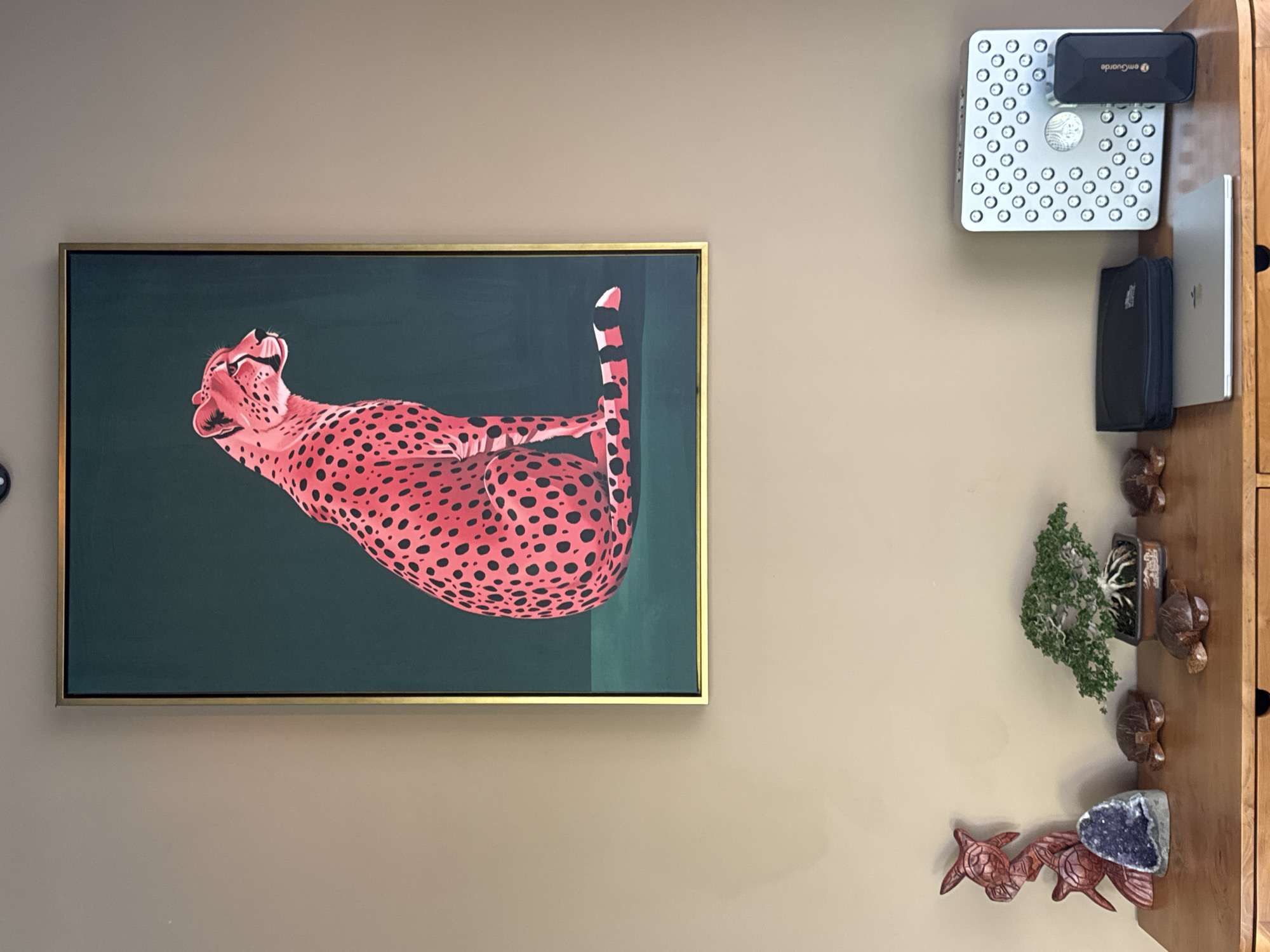 A room in a building in Kuta Selatan, Indonesia is depicted. A framed painting of a pink cheetah against a dark green background is centered on the left wall. A table stands against the right wall with a silver device, a black device, a silver laptop, a black case, a bonsai tree, carved wooden turtles, and an amethyst geode sitting on it. The walls are painted a neutral beige color.
