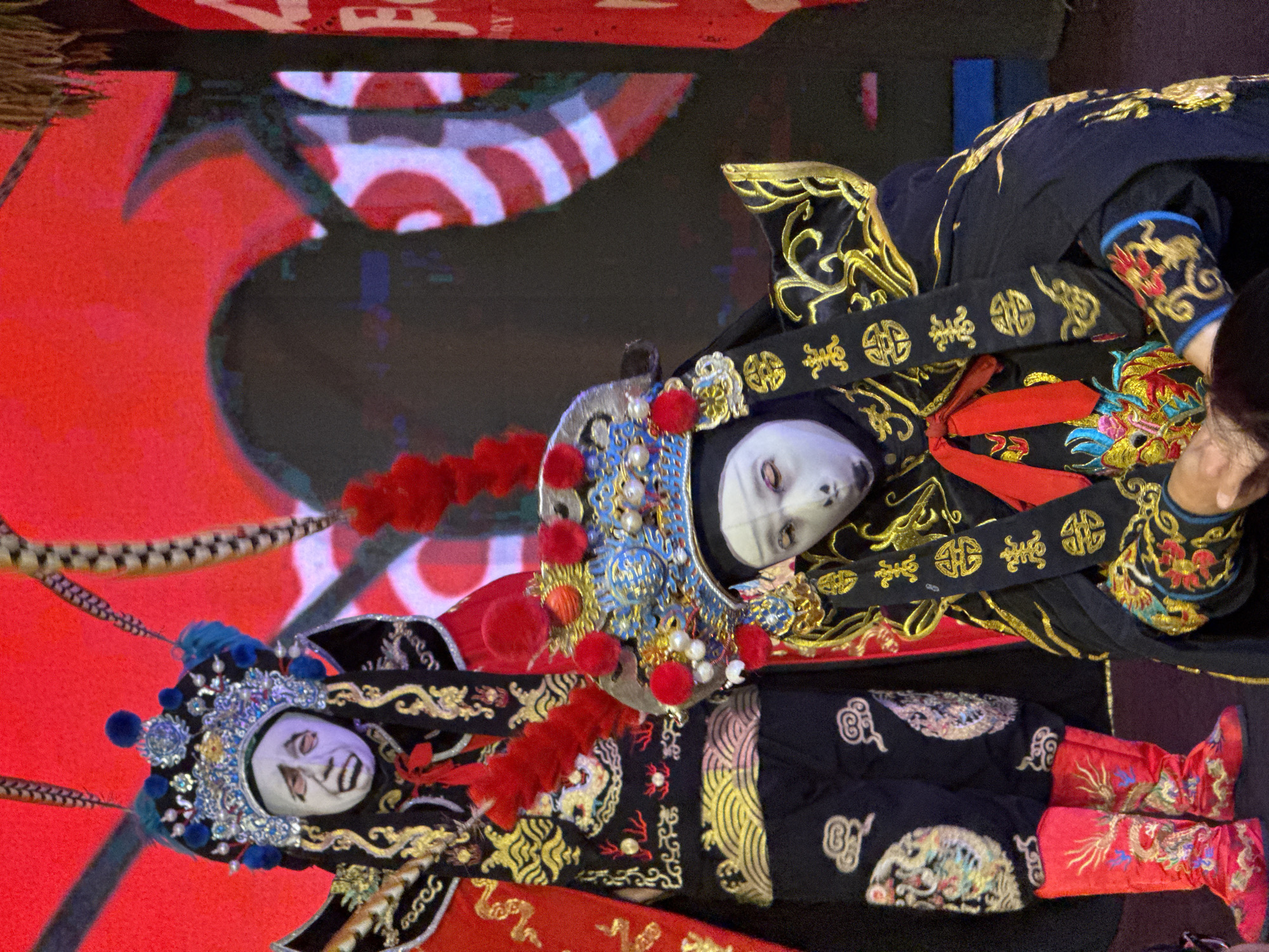 The image displays two figures, likely performers or large puppets, adorned in elaborate traditional Chinese costumes within a theatrical setting. The photograph is oriented vertically (rotated 90 degrees clockwise).
The subjects are dressed in opulent black and gold attire, richly embroidered with intricate patterns. Their headpieces are a focal point, featuring detailed metallic designs, jewels, and vibrant blue and red pom-poms; one headpiece also includes prominent feather-like extensions. Both figures exhibit stylized white faces with painted black eyes and mouths, characteristic of Chinese opera masks or highly theatrical makeup.
The costumes feature broad sashes or belts with repeating gold embroidered stylized Chinese characters, specifically resembling "福" (fú), which symbolizes good fortune or blessing. One figure wears striking red boots, also adorned with gold embroidered motifs, possibly depicting dragons or other auspicious creatures.
The background consists of a brightly lit, dynamic screen, predominantly red with abstract black and white patterns, suggesting a modern stage backdrop or LED display. The setting appears to be an indoor performance venue, possibly in Yuzhong District, China, aligning with the cultural attire. The activity depicted is likely a performance, with the figures in a static, posed moment that highlights the grandeur and artistry of their traditional garments. Artificial stage lighting illuminates the scene, enhancing the vibrant colors and intricate details. No specific time of day or weather conditions are discernible.