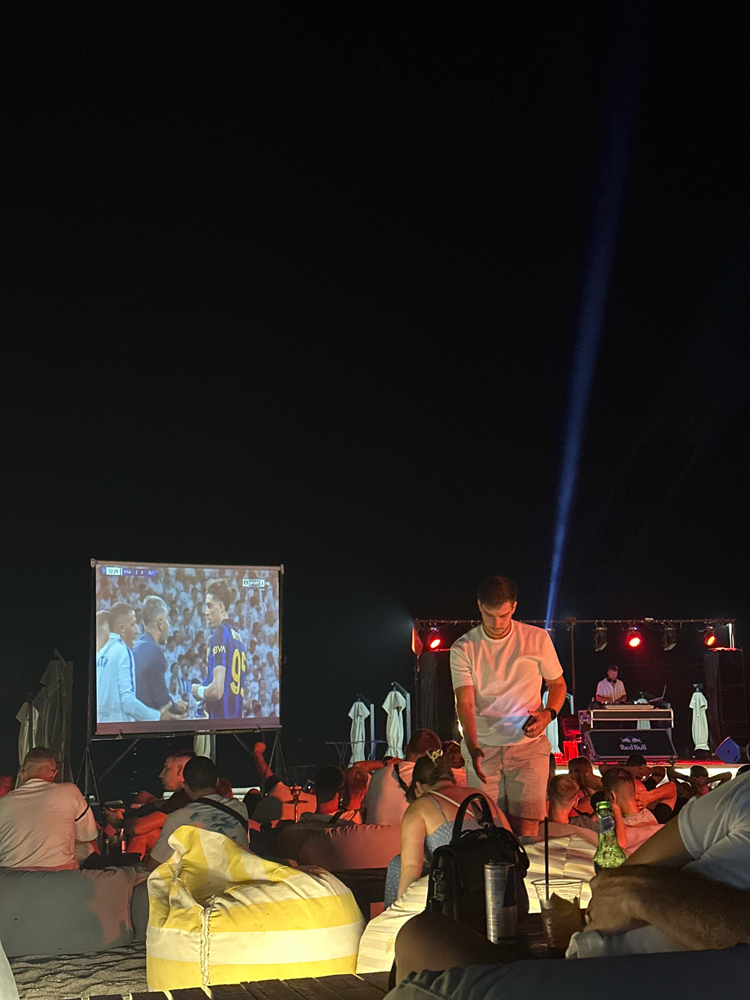 A nighttime outdoor gathering is taking place, with a large projector screen displaying a soccer match. Several people are seated on beanbag chairs, facing the screen. In the foreground, a man in a white t-shirt and light-colored shorts stands near a table with drinks. Behind him, on a stage with red lighting and a blue spotlight, a DJ is set up. A crowd of people is visible in the background, watching the match or the performance. The environment appears to be a beach or outdoor venue, with some structures and umbrellas visible in the distance.