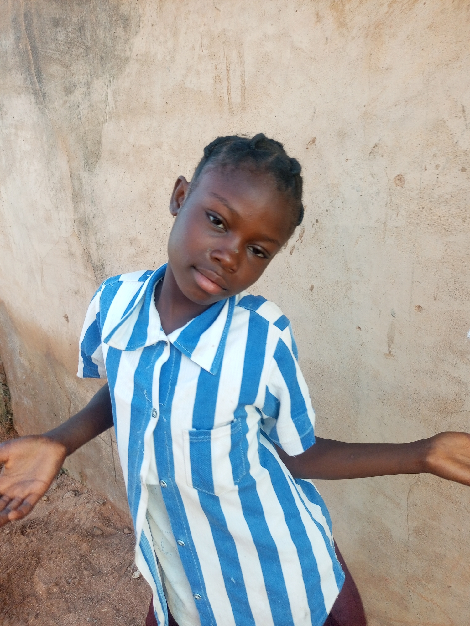 A young girl with dark skin and braided hair stands in front of a rough, beige wall. She is wearing a blue and white striped button-down shirt with a collar. Her hands are extended outwards, palms facing upwards, in a questioning gesture. The ground beneath her appears to be reddish-brown dirt. The scene is outdoors, likely in a rural or less developed area.