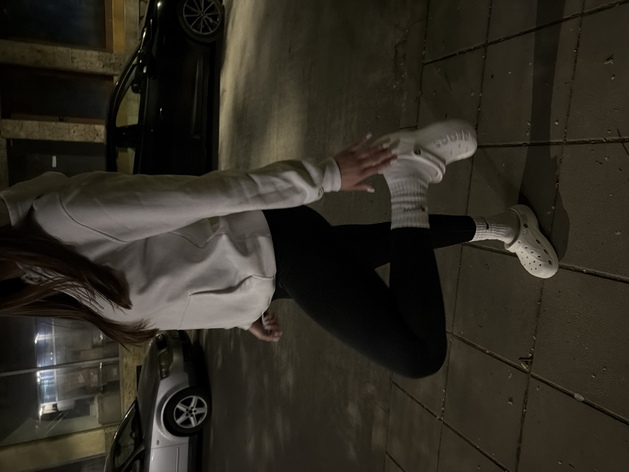 A person, viewed from above and to the side, stands on a tiled walkway. They wear black leggings, a light gray sweatshirt, and white Crocs over white socks. One hand is visible, with white nail polish. To the left of the person are two parked cars, one black and one silver. The environment suggests a street or parking area at night or in low light. The city of Lovech, Bulgaria, is not discernible from the image.
