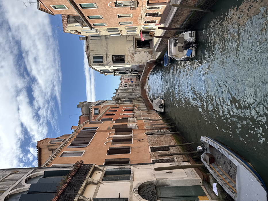The image presents a narrow canal in Venice, Italy, framed by multi-story buildings under a partially cloudy blue sky. On the left bank, a light-colored building featuring a large arched dark green door and an upper-story balcony with dark shutters stands in the foreground, adjacent to a reddish-orange building with numerous dark brown-shuttered windows. Further along the left, a white boat covered by a light tarpaulin with an outboard motor is moored beneath a reddish-brown arched stone bridge that spans the canal.
On the right bank, a reddish-orange building with light-shuttered windows and an ornate street lamp is visible, followed by a lighter-colored building displaying cracked plaster, dark shutters, and a ground-level entrance with a red awning.
Within the greenish-brown canal water, a long, light grey utility boat with a small outboard motor is moored on the left in the foreground. On the right, a white recreational motorboat with an outboard motor and two visible seats is docked, positioned in front of a partially visible smaller dark blue boat. No individuals are present in the scene.