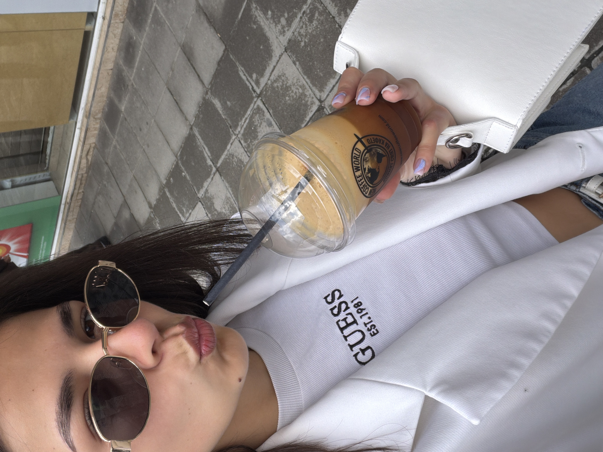 A female is shown in a close-up selfie, holding a cup of iced coffee while wearing sunglasses. The coffee cup features a logo for "WE WORLD COFFEE - СВЯТ НА КАФЕТО", identifying it as a location within the city of Gabrovo, Bulgaria. The woman is dressed in a white top with "GUESS EST. 1981" printed on it, paired with a white blazer and a white purse. The background shows an urban setting with tiled pavement and a glimpse of greenery on the side. The woman is posing with her lips puckered.