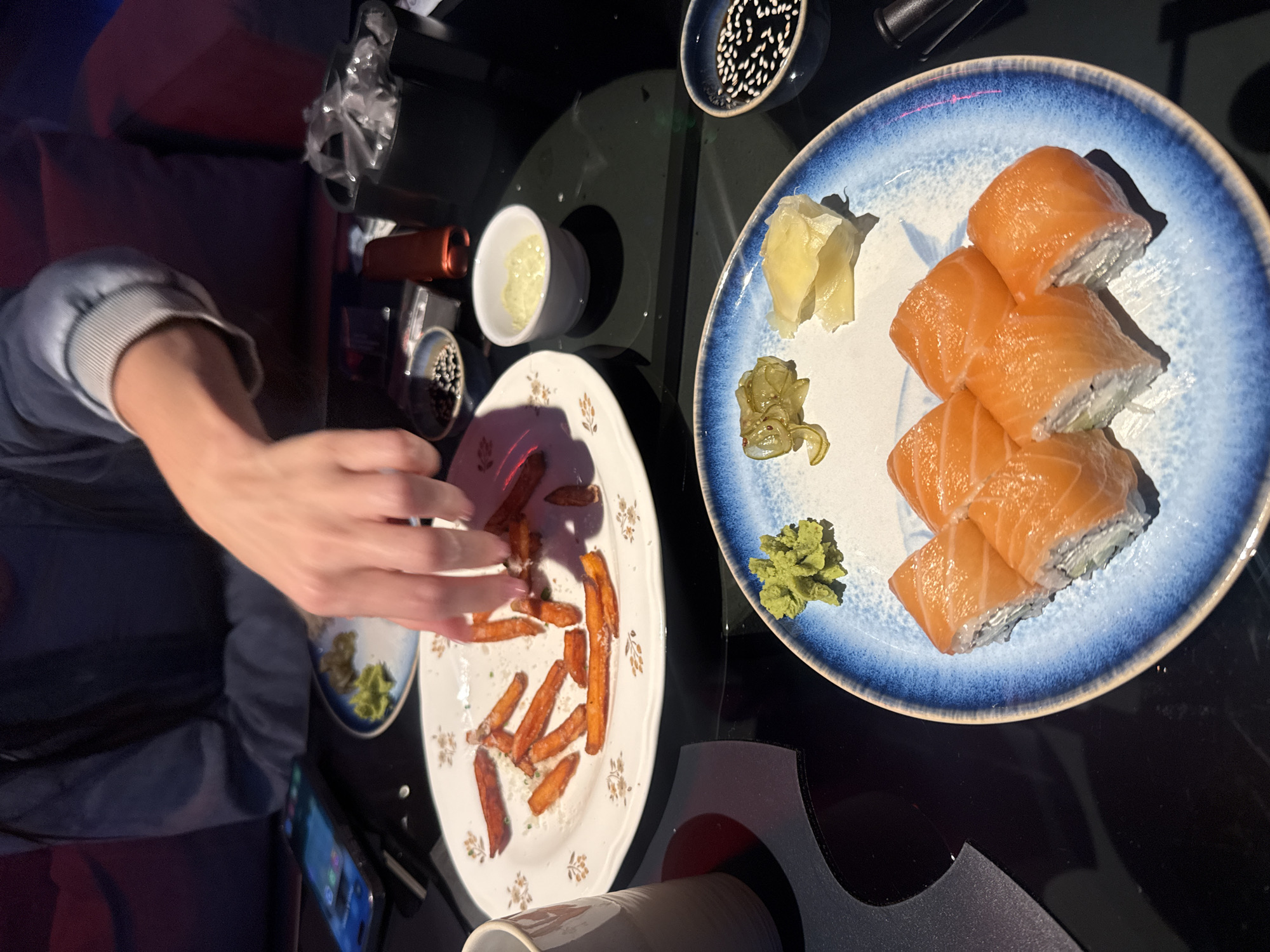 The scene depicts a table setting with plates of food, likely in an indoor restaurant or lounge. A person's hand is reaching towards a plate of what appears to be sweet potato fries. Another plate holds sushi rolls with salmon, accompanied by wasabi and pickled ginger. Small bowls contain dipping sauces and sesame seeds. A smartphone is visible on the table beside the plates. The location cannot be confidently inferred from the provided image without further information. Moscow, Russia can not be inferred from the picture.