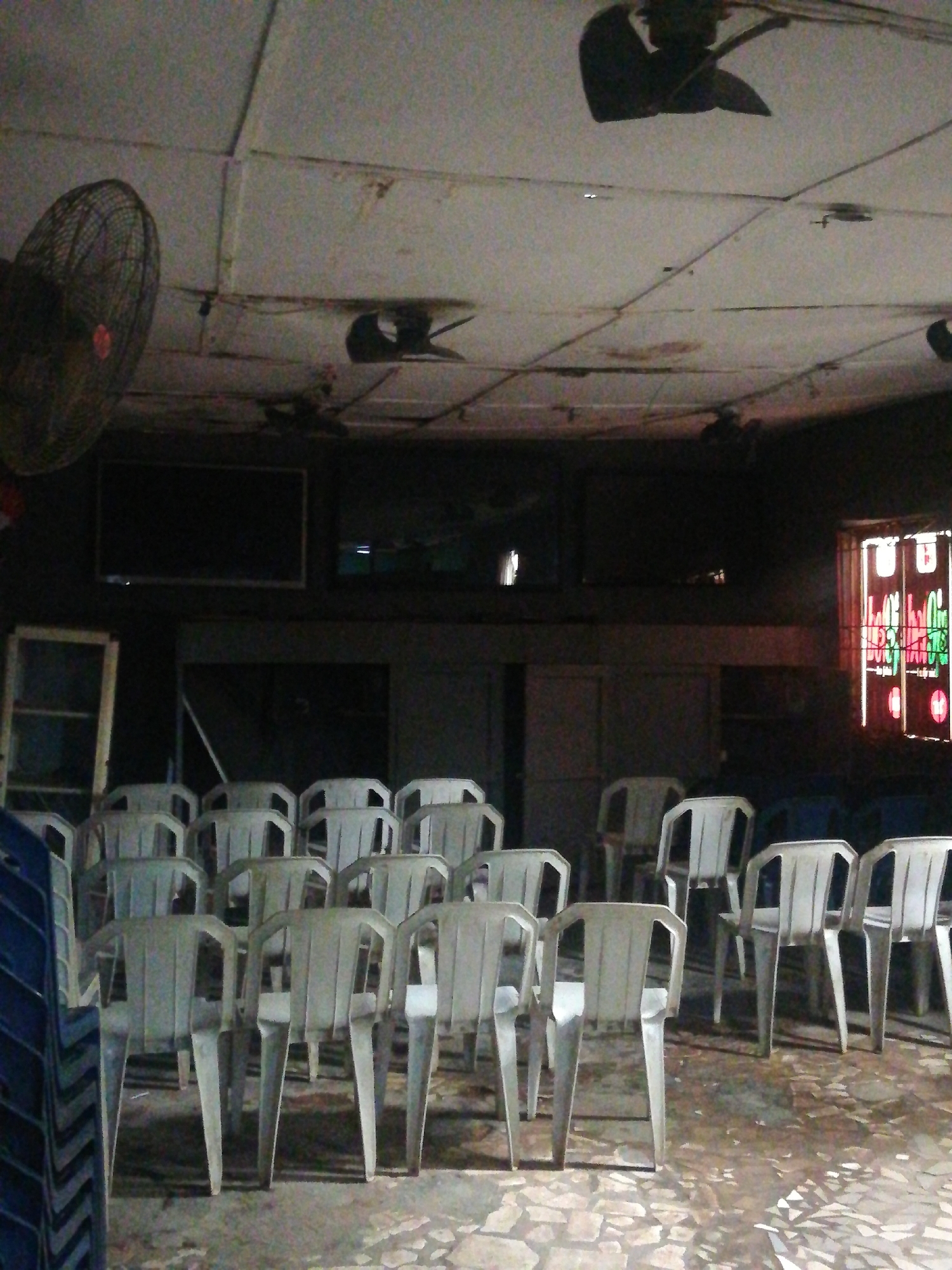 The image depicts an interior space, likely in a building in Lagos, Nigeria. Rows of white plastic chairs face a dark wall where three large televisions are mounted. A window with decorative glass panels is visible on the right. A large, old-fashioned fan is mounted on the wall to the left. The ceiling appears damaged with dark, irregular shapes and what seems to be electrical wiring exposed. The floor has an uneven, tiled surface. Stacks of blue plastic chairs are visible on the left. The overall lighting is dim. There are no people visible in the image.