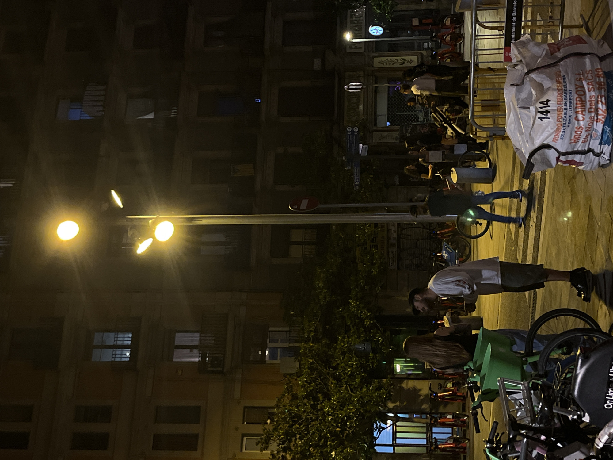 Captured at night in Barcelona, Spain, the image reveals an active urban street scene. Three illuminated streetlights cast a glow on a multi-story building facade. A group of people are gathered near a bicycle parking station, engaging in conversations and standing in close proximity. The bicycles, along with additional bikes and scooters, occupy the foreground. Notably, a large, white industrial sack labelled with "PROHIBIDO PONER FIBROCEMENTO SOLO UTILIZABLE EN BARCELONA" is visible, partially obstructed by metal fencing. The text indicates the prohibited use of asbestos-cement in Barcelona. A sign indicating "Juntament de Barcelona" further confirms the location.