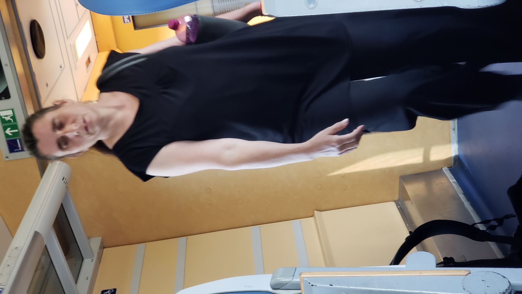 The image shows a woman standing inside what appears to be a train car. She is wearing a black sleeveless top and black pants, and is holding a purple water bottle. The train car has yellow walls with circular decorative elements, and a blue floor. There are signs on the wall above the woman, including one with a wheelchair symbol and another with arrows indicating direction and an exit. In the foreground, a backpack with black straps is visible. The lighting suggests it is daytime, with light coming from above and from the right side of the image.