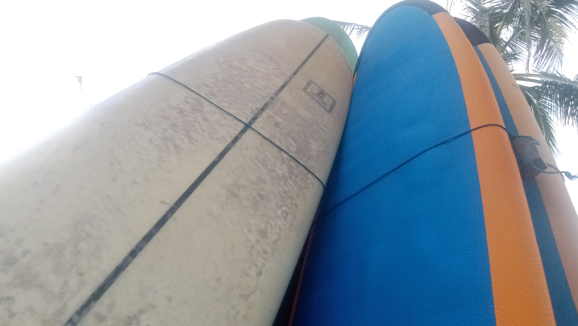 Two surfboards, one light-colored and weathered, the other a vibrant blue and orange, lean against each other. They are positioned outdoors, with a hint of palm fronds visible in the upper right corner, suggesting a tropical location like Kuta, Indonesia. The sky above is bright and overcast. The surfboards appear to be resting, possibly stored or waiting for use. There are no people or animals in the frame, and no discernible activity is taking place. The focus is solely on the surfboards and their immediate surroundings.