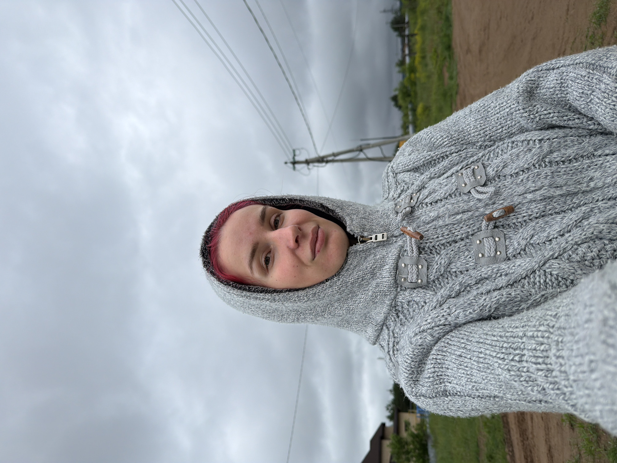 The image captures a selfie of a female individual with short, reddish-pink hair, wearing a light gray, cable-knit hooded cardigan with the hood up, set against an overcast sky in Chaikovsky, Russia. The individual, positioned slightly right of center and looking directly at the camera, has fair skin and a neutral expression. The cardigan features a zipper and three prominent toggle closures with wooden toggles and grey faux-leather or leather straps adorned with small metal studs.
The background reveals an undeveloped or semi-rural setting under a heavily clouded, gray sky. To the far left, a modern-style house with a brown roof is partially visible amidst green foliage. In the middle-ground to the right, a wooden utility pole with multiple overhead power lines extends diagonally upwards, with the lines running horizontally across the upper right quadrant of the image. The ground consists of a dirt path or road in the foreground and right, with patches of green grass and scattered low-lying vegetation. Distant trees and faint structures are visible along the horizon line beneath the dense clouds.