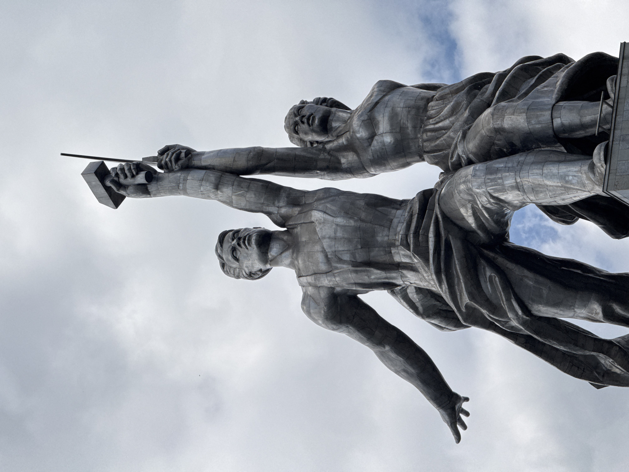 The image shows a monumental metal sculpture of two figures against a cloudy sky. One figure stands atop the other, both with their arms raised high. The figure on top holds a hammer and the figure on the bottom holds a sickle. The sculpture is part of the "Worker and Kolkhoz Woman" monument by Vera Mukhina. The statue is located in Moscow, Russia, at the VDNKh (Exhibition of Achievements of National Economy).