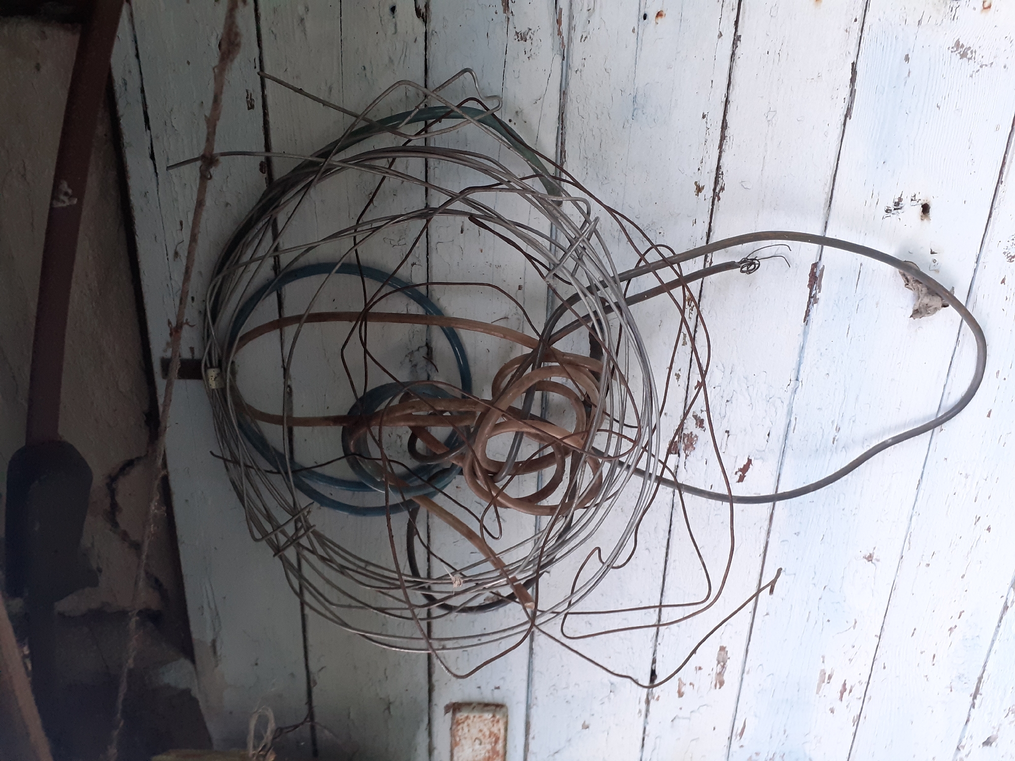 An outdoor scene in Deremezna, Ukraine, features a weathered wooden wall with peeling white paint. A tangled mass of various wires and cables, some appearing rusty and others insulated in brown and blue, hangs suspended from a horizontal rope attached to the wall. A small, rusted metal plate is visible on the wall to the left of the hanging wires. The overall impression is one of disuse and storage of electrical components.