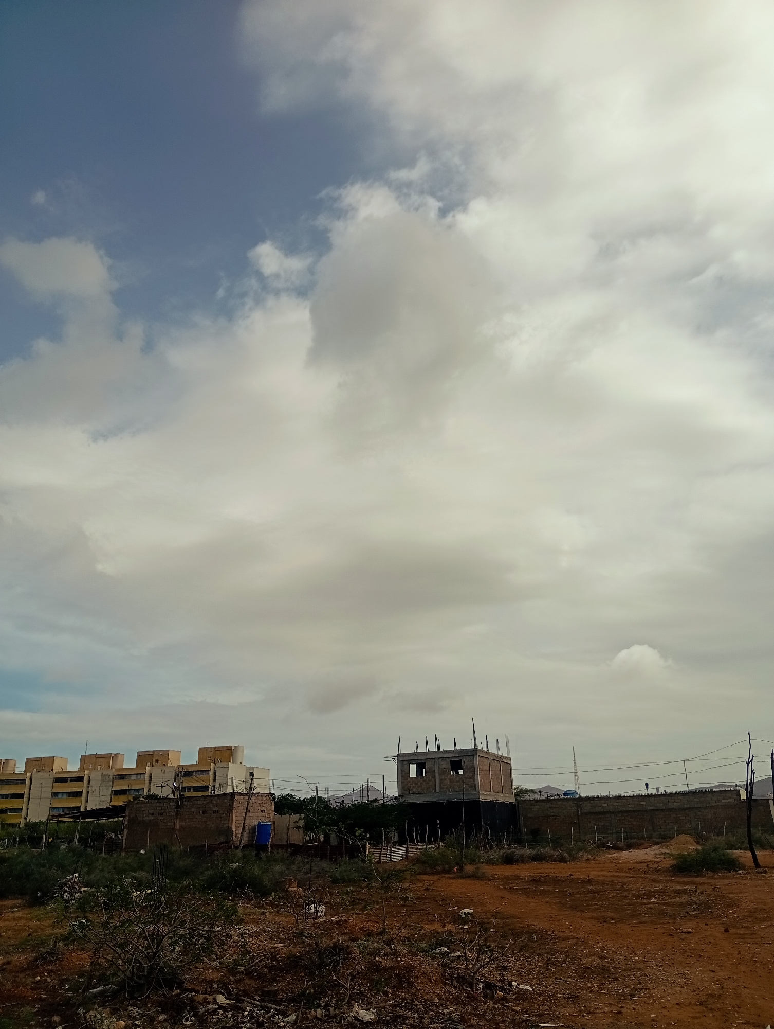 The image depicts a cloudy sky over a semi-arid landscape with several buildings under construction and some completed structures. In the background, a row of light-colored, multi-story apartment buildings with visible windows are present. In the mid-ground, a walled construction site features a partially built, two-story concrete structure with visible rebar, suggesting ongoing construction. Adjacent to this is a lower, brick-built structure with a blue water tank on its roof, and various makeshift shelters or sheds are scattered around the area. The foreground consists of dry, reddish-brown soil with sparse vegetation, including dry bushes and debris. The scene suggests an urban or semi-urban environment undergoing development, with a mix of residential and ongoing construction projects. The location is Altagracia, Venezuela.