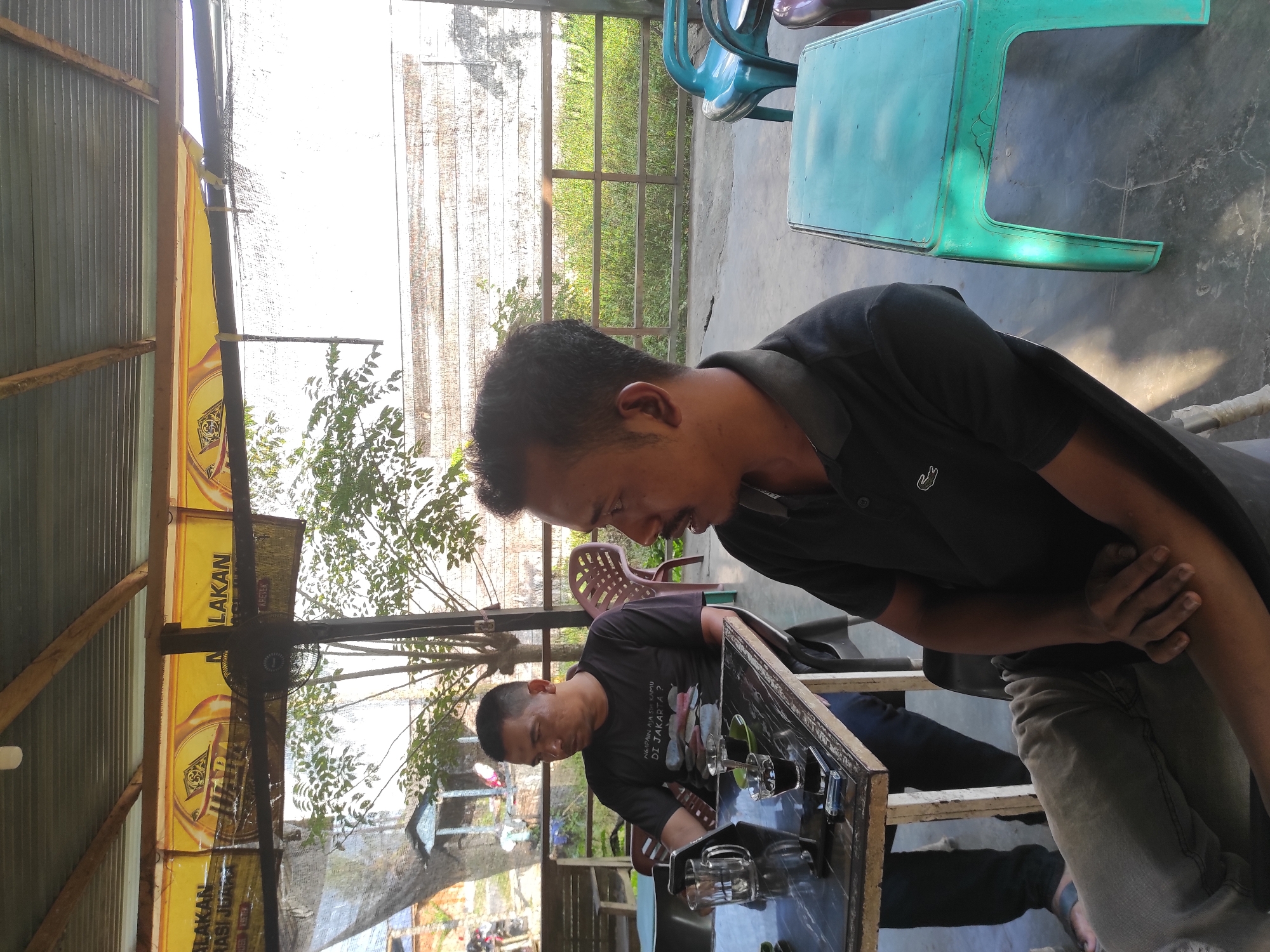 Two men are sitting at a table outdoors. One man is wearing a dark polo shirt with a logo. The other man is wearing a black t-shirt with a graphic. The table is covered with glassware. A turquoise plastic chair is nearby. A partial view of a fence and trees are in the background. The scene is in Lhokseumawe, Indonesia.