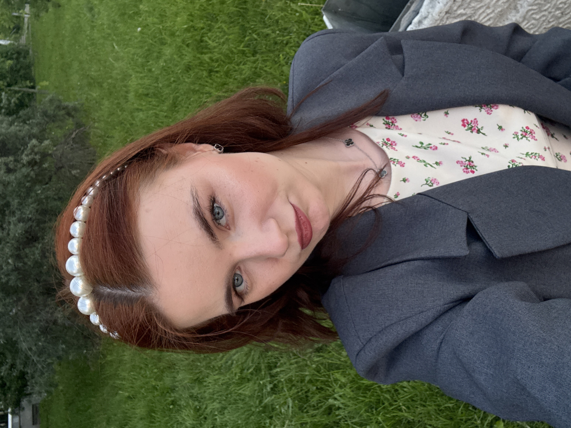A young woman is depicted in a close-up portrait, oriented in a vertical format, with the image rotated 90 degrees counter-clockwise. She looks directly at the camera with a slight, gentle smile, her head tilted. Her reddish-brown hair is adorned with a white pearl-beaded headband. She has striking blue-green eyes, defined eyebrows, and wears dark red lipstick.
She is dressed in a dark grey blazer worn over a light-colored top featuring a delicate floral pattern with small pink and green flowers. A silver necklace with multiple small, dark, clover-shaped charms is visible around her neck, and a small earring is in her visible ear.
The background primarily shows vibrant green grass, suggesting an outdoor setting like a park or a garden. Some faint trees or bushes can be seen in the distance on the left side of the frame. The lighting indicates it is daytime, with soft, natural light. No specific activity is taking place other than posing for the photograph. The overall impression is calm and pleasant. The context for this scene is Saint Petersburg, Russia. There is no visible text in the image.