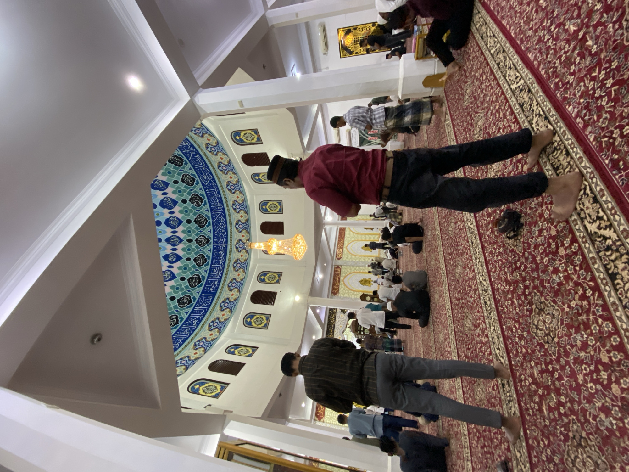 The image depicts the interior of a mosque in Lhokseumawe, Indonesia. The mosque features a large, ornate dome with Islamic calligraphy and intricate tilework in shades of blue, green, and white. A large, multi-tiered chandelier hangs from the center of the dome. The floor is covered with a red, intricately patterned carpet. Several people are present, most of whom are men dressed in traditional attire, including sarongs and shirts. Some are standing, while others are seated on the carpet, appearing to be engaged in prayer or religious activities. In the foreground, two men are standing, facing away from the camera, their backs to the viewer. The overall environment suggests a religious gathering.