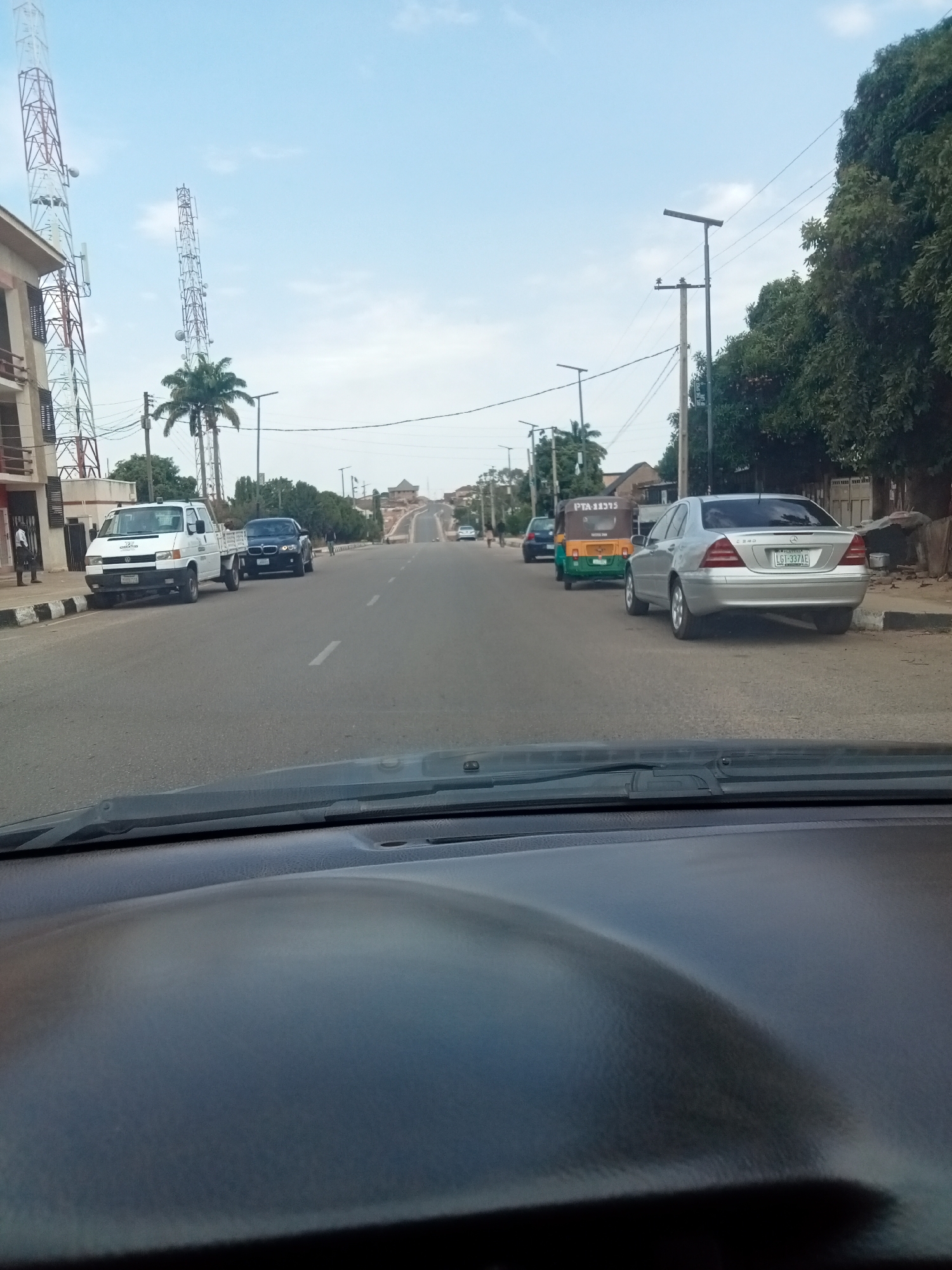 The image shows a view from inside a car, looking down a paved road in Jos, Nigeria. The road has multiple lanes and a median. On the left side of the road, there's a white van, a dark-colored car, and a building with telecommunication towers. On the right side, there is a parked silver Mercedes-Benz, a green and yellow tricycle (keke), and trees. Overhead power lines are visible. The sky is partly cloudy.