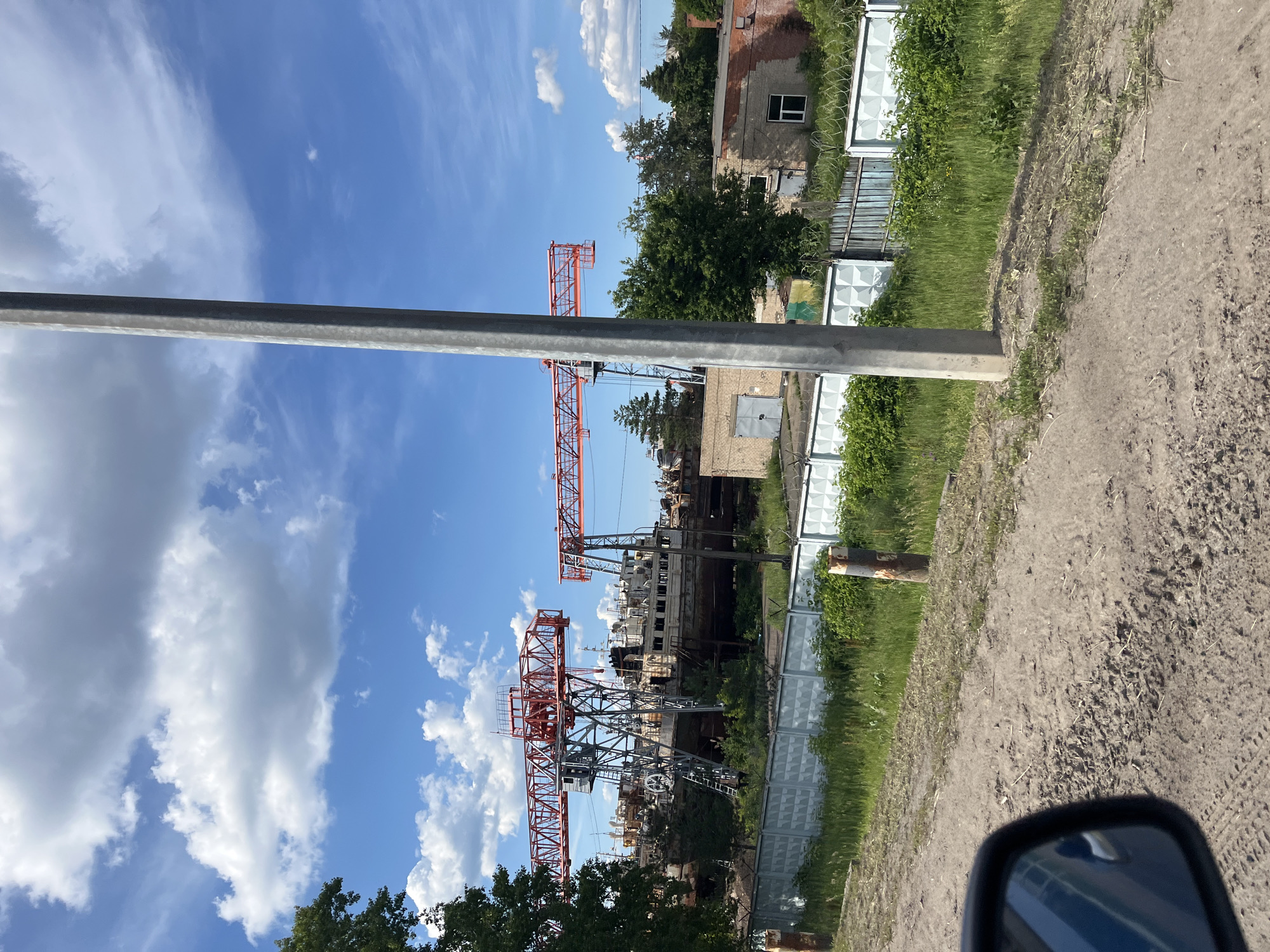 The image shows a construction site under a bright blue sky with scattered white clouds, indicating daytime and fair weather. The scene is dominated by tall red construction cranes, suggesting ongoing building or industrial activity. In the foreground, a grey pole or pillar is visible, possibly part of the infrastructure or a street light.
Behind the cranes, several brick buildings with windows and doors are present, some appearing weathered. A fence, made of white patterned panels and wire mesh, runs along a portion of the site. Lush green grass and trees surround the buildings and fence, indicating a temperate climate. The ground in the foreground appears to be a dirt or gravel road, with tire tracks suggesting vehicle activity. In the lower right corner, a car's side mirror is visible, reflecting the sky and a portion of the road.
The overall impression is one of an active industrial or construction area in a semi-urban or rural setting. There is no visible text in the image. The orientation of the image is vertical, with the sky appearing at the top and the ground at the bottom.