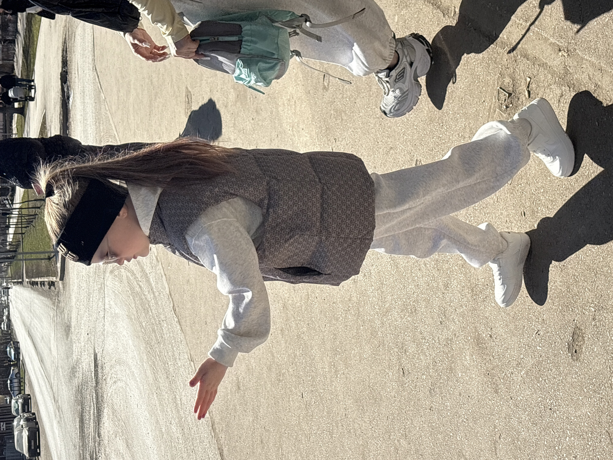 A young girl, seen from the chest up, stands on a sidewalk. She wears a black headband, a gray patterned vest, and a light gray sweatshirt and sweatpants, along with white sneakers. She extends her right hand downwards. To the left, a street with parked cars lines the road. Two more people are walking ahead of the girl, also wearing sweats and sneakers. One is wearing a backpack. The location is the city of Perm, Russia.