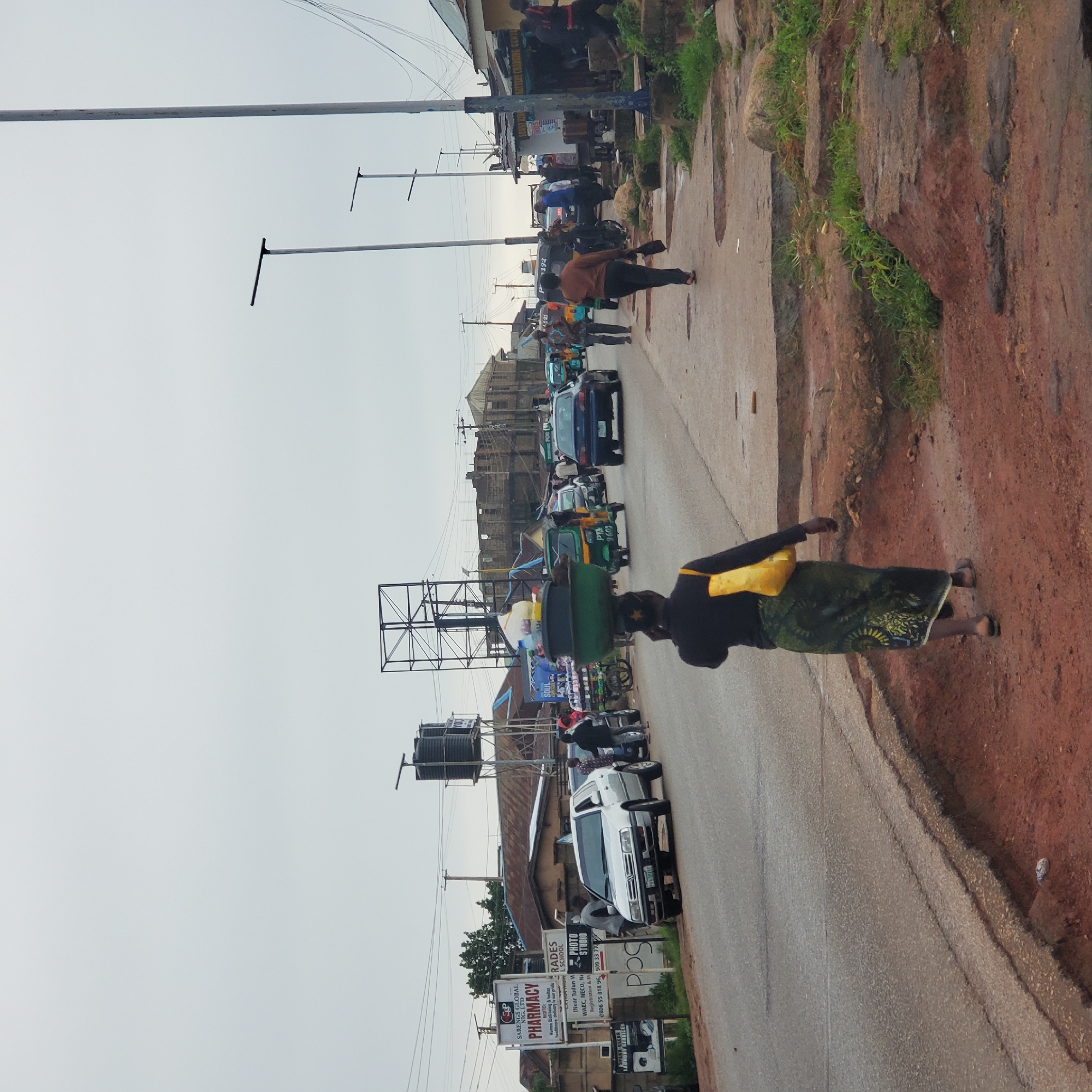 In the city of Jos, Nigeria, on a cloudy day, a street scene unfolds with a mix of pedestrian and vehicular activity. In the foreground, a woman walks away from the camera, carrying a large green basin filled with items on her head. She is dressed in a dark top and a patterned wrap skirt, with a yellow bag slung over her shoulder. The road surface is paved but shows signs of wear and tear, with some erosion visible on the roadside.
Vehicles are present on the road, including a silver sedan, a blue sedan, and several motorized tricycles, commonly known as "keke-napeps". People are seen standing and walking along the roadside, some near shops and stalls. Buildings line the street, with some appearing to be commercial establishments, indicated by signage for "Pharmacy", "Photo Studio", and "Laundry Services". A water tower with large black tanks is visible in the background, along with various utility poles and wires. The overall environment appears to be a bustling urban street with signs of everyday commerce and transportation.