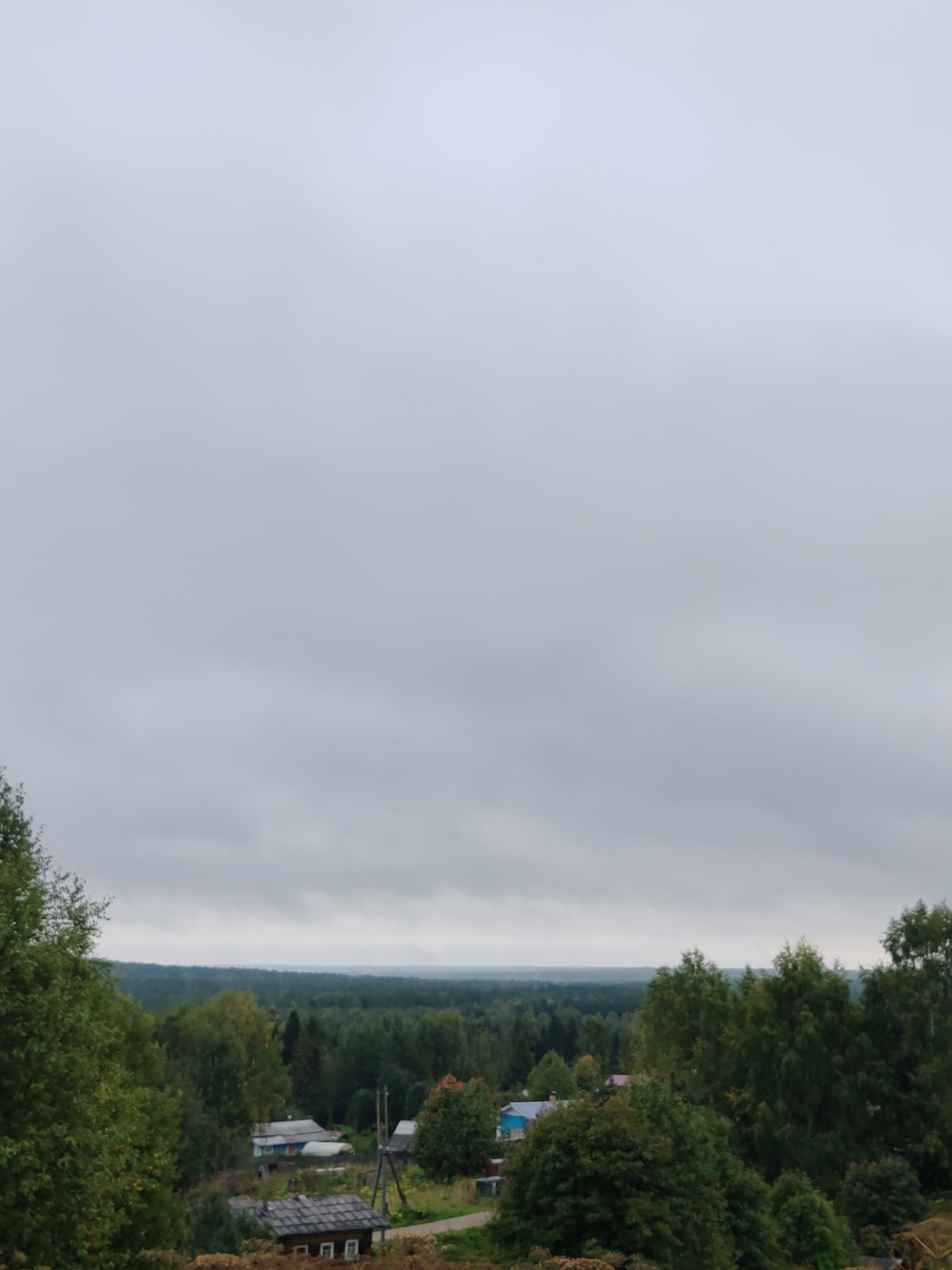 The image captures an overcast sky above a rural landscape in the Chuchlam, Russia region. In the foreground, lush green trees dominate the left side of the frame, with some foliage extending into the lower right corner. Below the trees, a small collection of houses is visible. One house has a gray roof and blue and yellow accents. Another house, situated closer to the foreground, has a dark gray roof and appears to be a single-story structure with visible windows. A wooden utility pole with some wires is also present near the houses. The middle ground is filled with a dense forest of various shades of green, stretching towards the horizon. The background shows a faint line of trees under a vast, gray sky with diffused clouds.