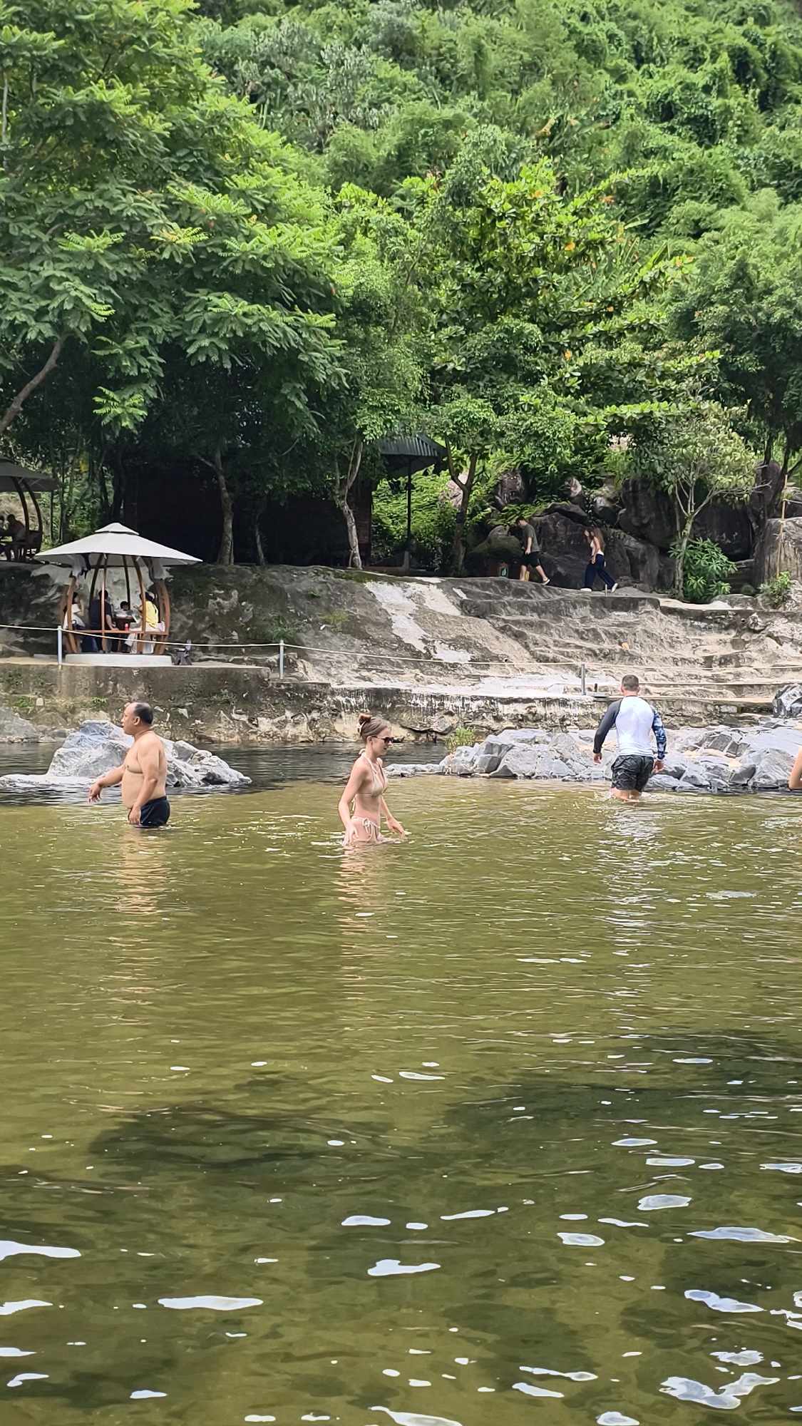 This image depicts a natural swimming area with a river or stream flowing through a lush, green environment. In the foreground, three individuals are wading in the water. On the left, a man in dark swim trunks is standing. In the center, a woman wearing a light-colored bikini is walking. On the right, a person in a long-sleeved rash guard and dark shorts is also in the water.
The background shows a rocky embankment leading up to a densely vegetated hillside. Several people are visible on the embankment and in the distance, some seated under a covered gazebo-like structure. The water is a murky, olive-green color, and ripples and light reflections are visible on its surface. The overall scene suggests a recreational spot in a natural setting. The location is identified as being in Khanh Vinh, Vietnam.