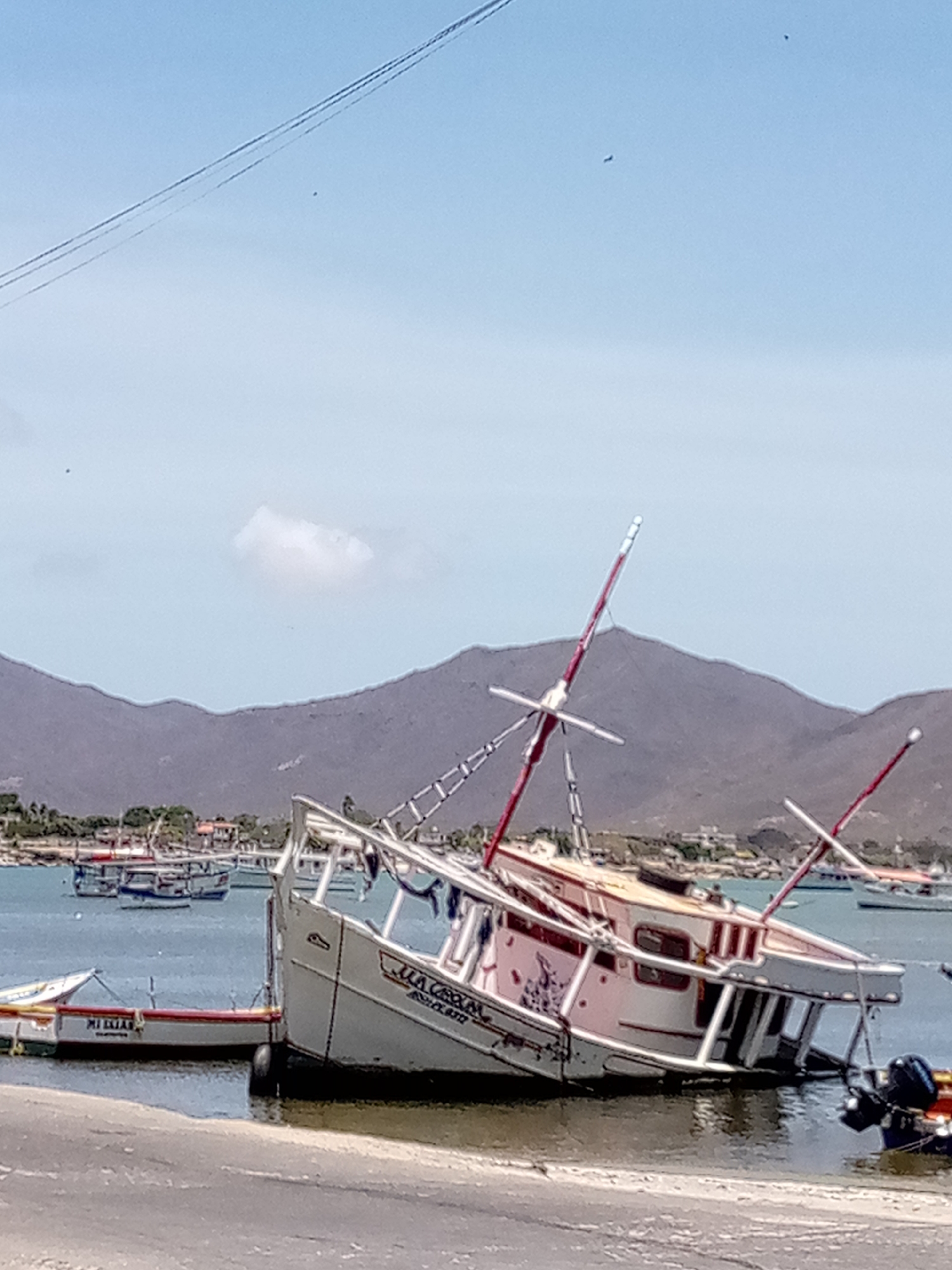 The image depicts a partially submerged, dilapidated boat on a shore. The boat is white and pink, leaning heavily to one side, with exposed interior framework and red masts. The name "La Ceona" is visible on its side. Smaller boats are docked nearby, including one labeled "Mi Elias". In the background, there is a bay with several more boats, a townscape, and mountainous terrain. The sky is partly cloudy. Power lines are faintly visible in the upper portion of the image. The location is identified as Juan Griego, Venezuela.