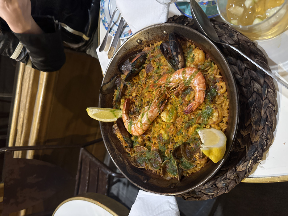 The scene is set indoors within a restaurant in Paris, France, at a dining table. The central subject is a large, dark metal paella pan filled with saffron-colored rice, featuring two prominent whole cooked prawns, numerous mussels and clams in their shells, scallops, and other seafood/meat pieces. The paella is garnished with a green herb sauce and two lemon wedges, one positioned upright at the top and another flat at the bottom.
The paella pan rests on a dark brown, circular woven placemat. To the left, part of a white table surface with a gold-colored rim is visible. To the right, a place setting includes a folded white napkin, two stacked white plates with a blue patterned rim, and a fork and knife. A clear glass containing an amber liquid with small white fruit pieces and a spoon is also present on the right side of the table.
In the background, a person is partially visible on the right side, wearing a dark jacket with light-colored horizontal stripes on the sleeve. Further back on the left, an empty, dark metal chair and another white table are discernible. The wall behind the table displays warm-toned paneling or molding. No explicit actions are depicted beyond the presentation of the meal.
